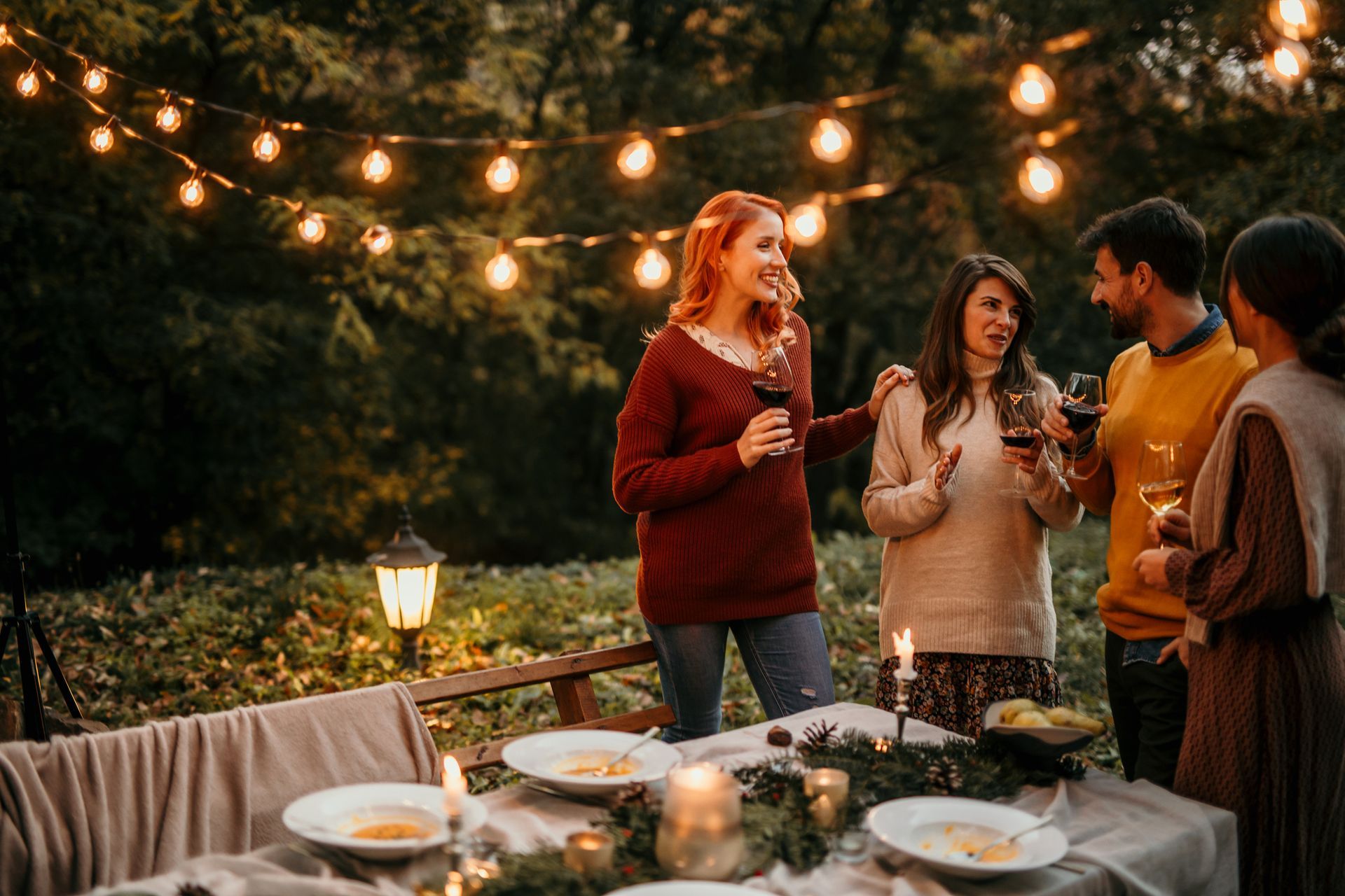 People at an outdoor dinner party, with string lights overhead.  Smiling, holding wine glasses, and gathering around a table.