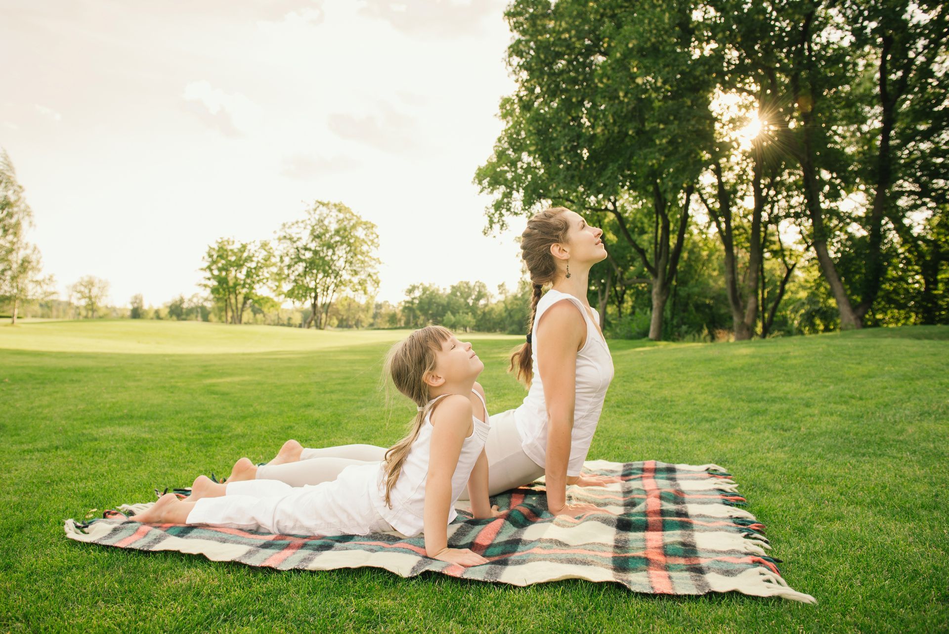 Woman and child in white outfits doing yoga on a blanket in a park.