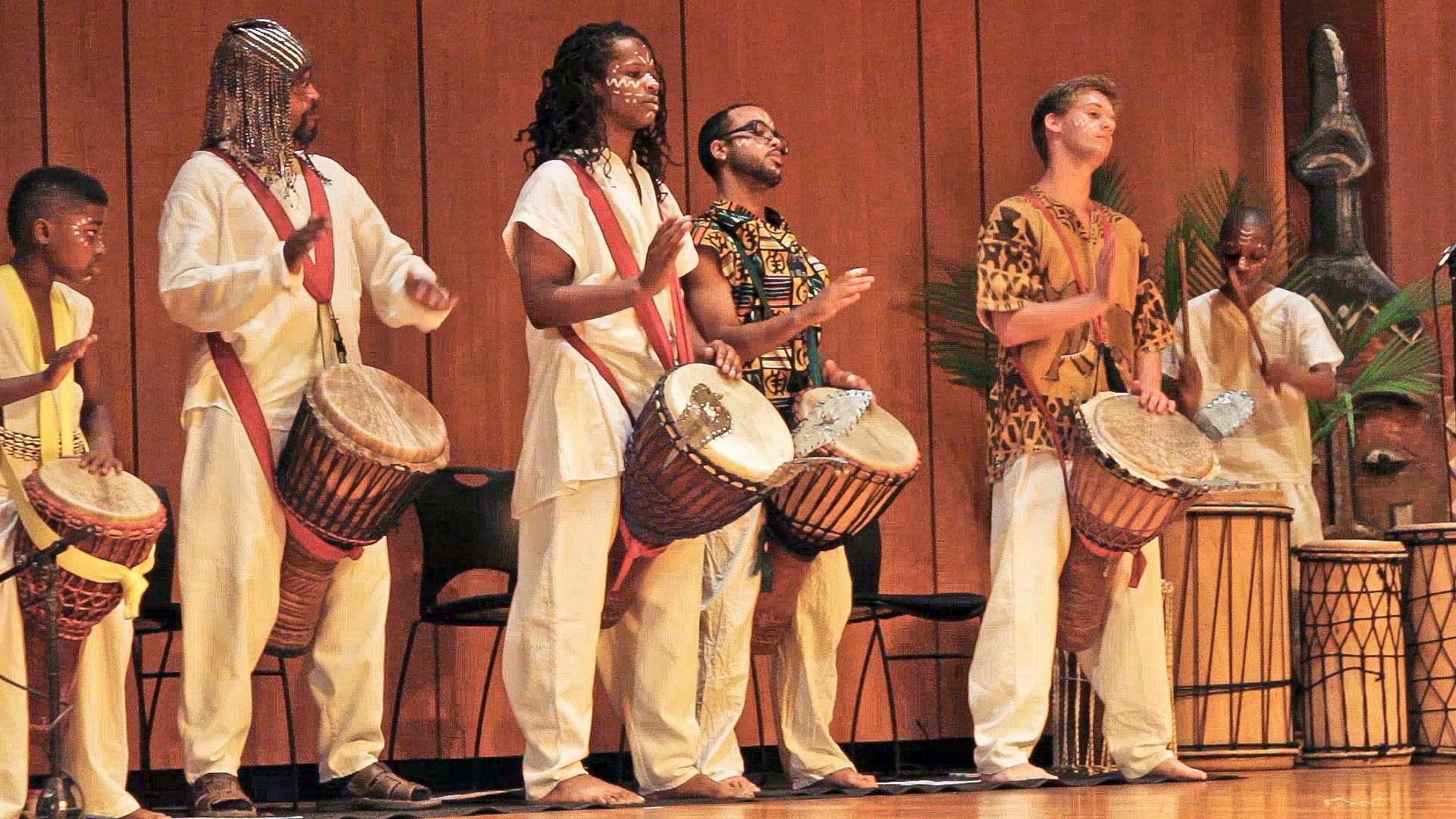 Group of drummers on stage, wearing white and patterned clothing, playing drums.