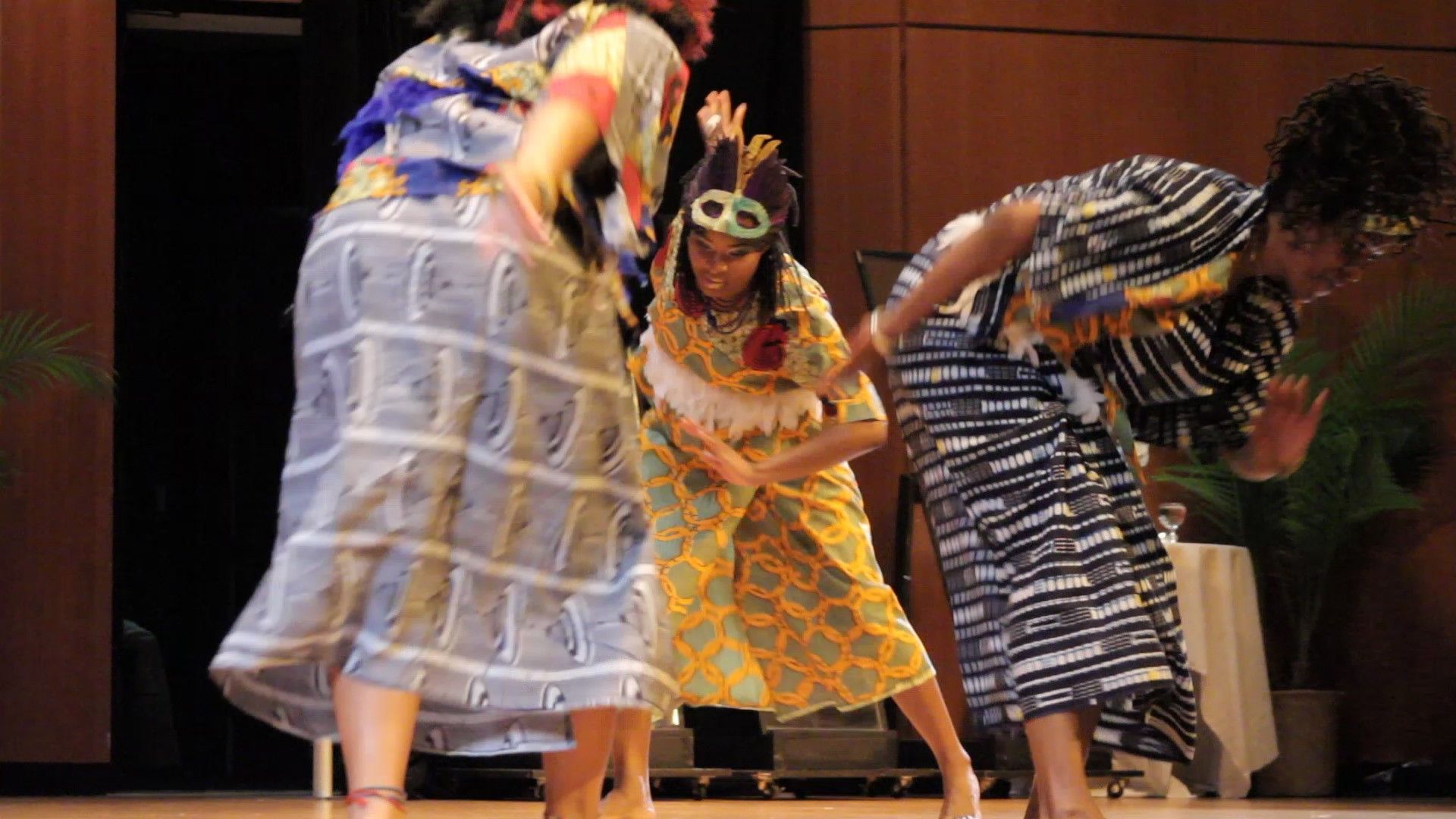 Three people dancing on a stage, wearing patterned dresses and smiling.