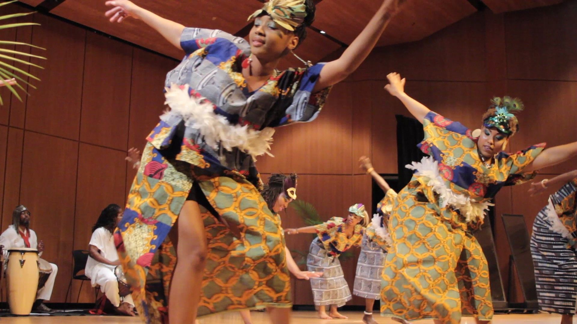 African dancers performing on stage, wearing colorful clothing, with musicians in the background.