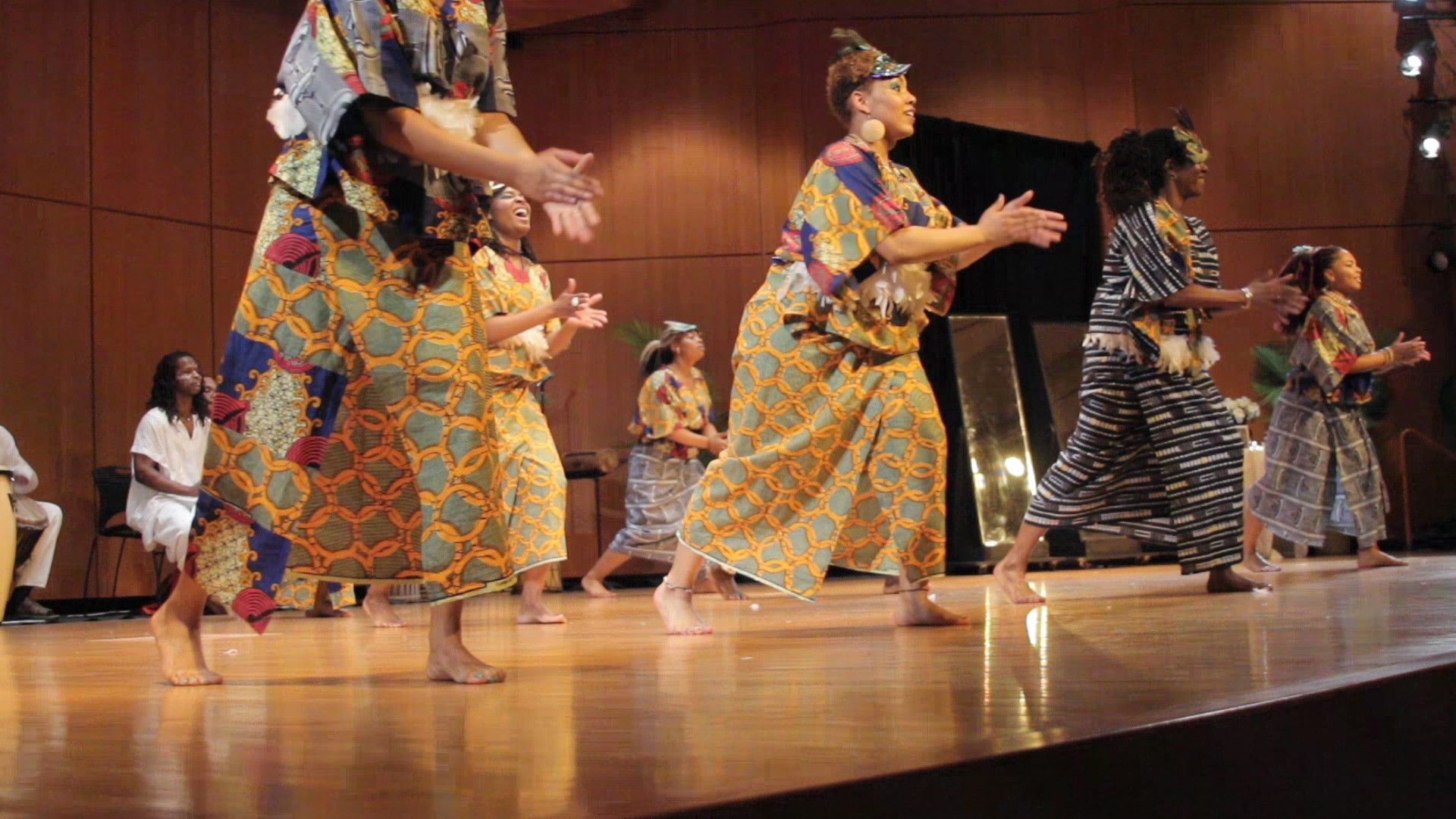 Group of women in colorful robes dancing on a stage, arms raised, indoors.