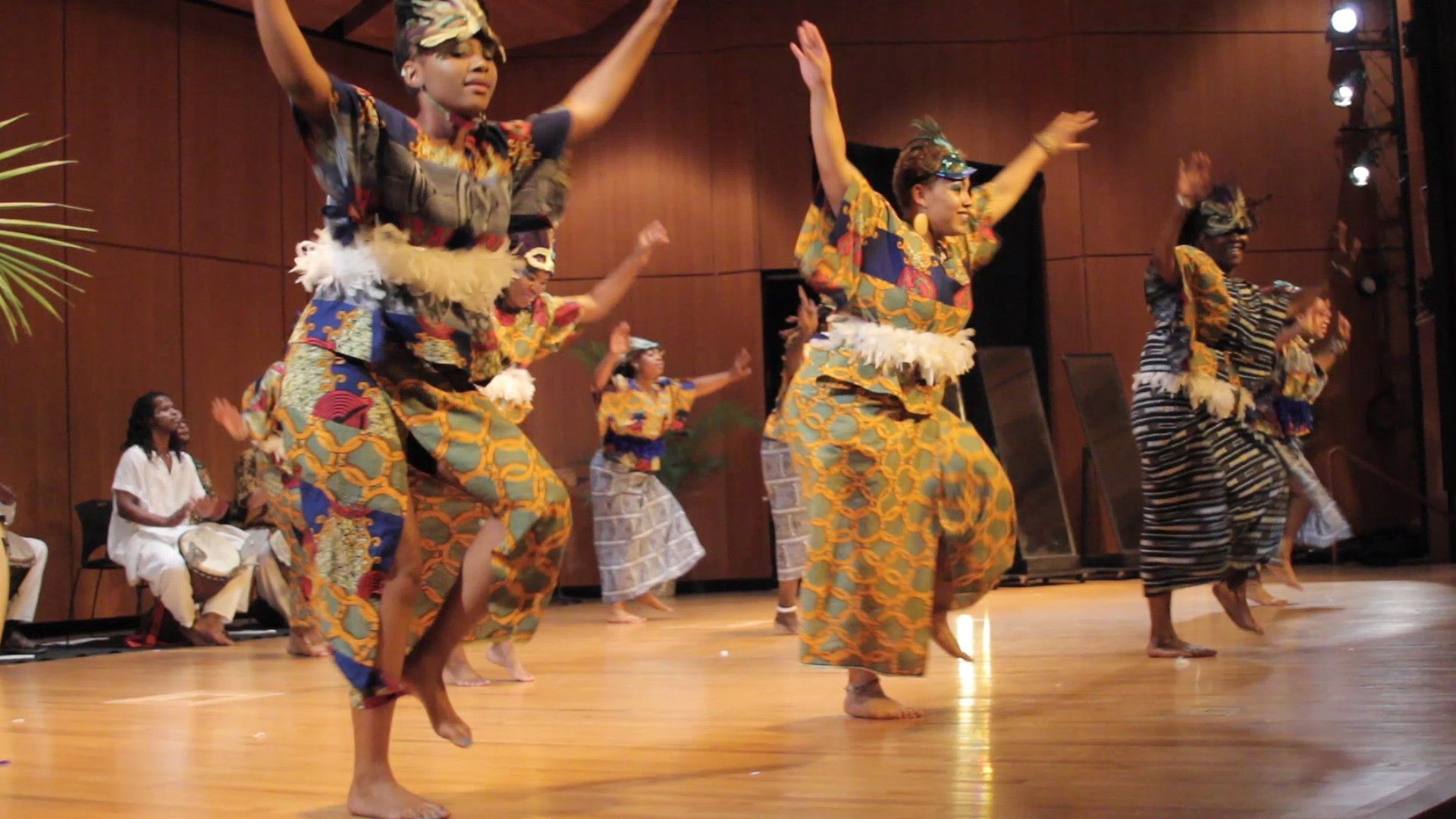 Dancers in colorful patterned costumes perform on a stage with arms raised.