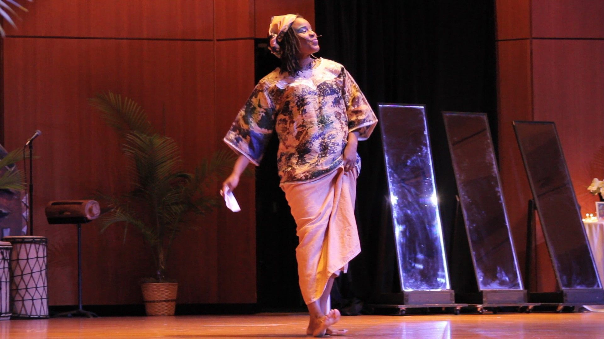 Woman in colorful clothing performing on stage; three lit rectangles behind her.