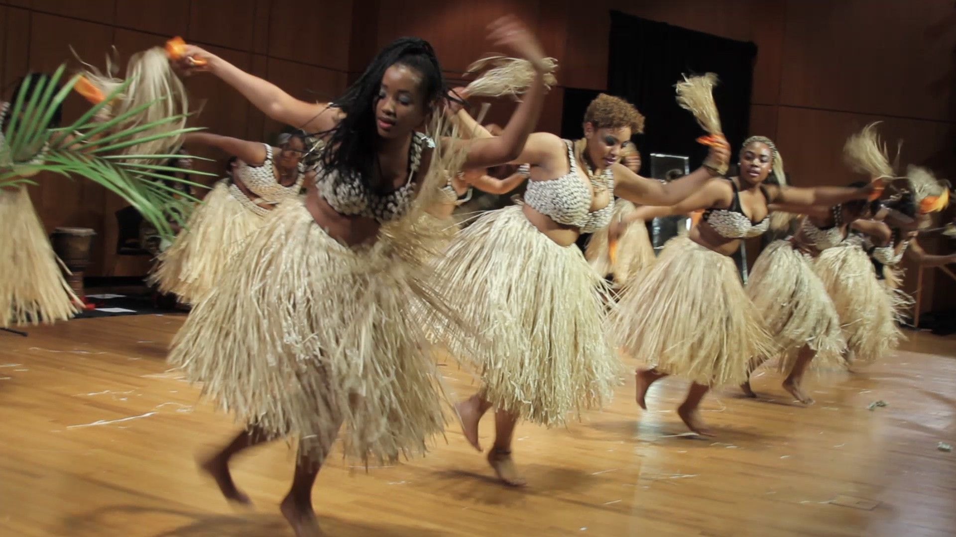 Group of Black women performing traditional dance with straw skirts and arm accessories on a wooden stage.