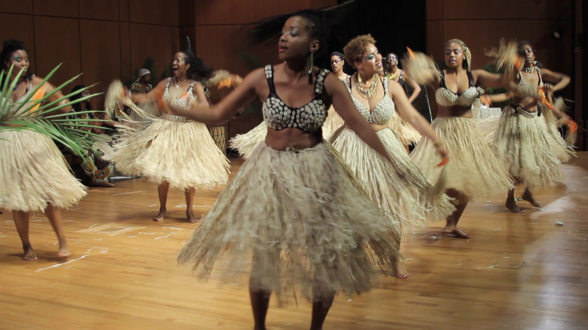 Dancers in straw skirts and tops perform on a wooden floor. They wave fans, smiling, in a large room.