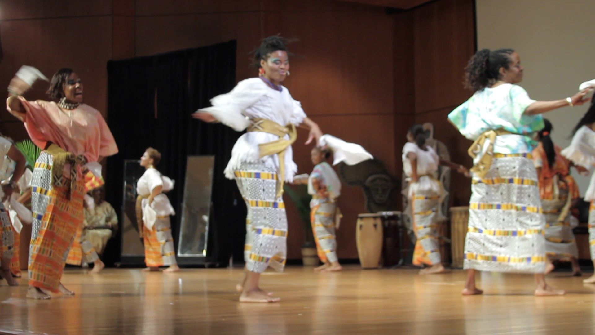 Group of women dancing on a stage in white and patterned skirts, some holding fans.