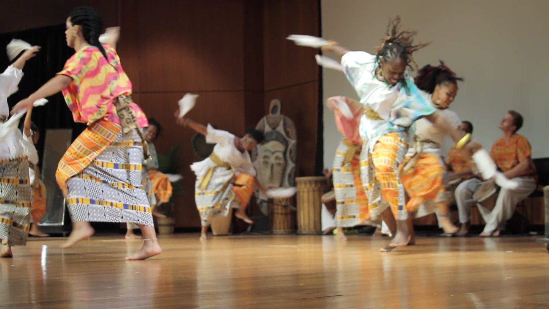 People dancing on stage in colorful clothing, holding white cloths, wooden floor.