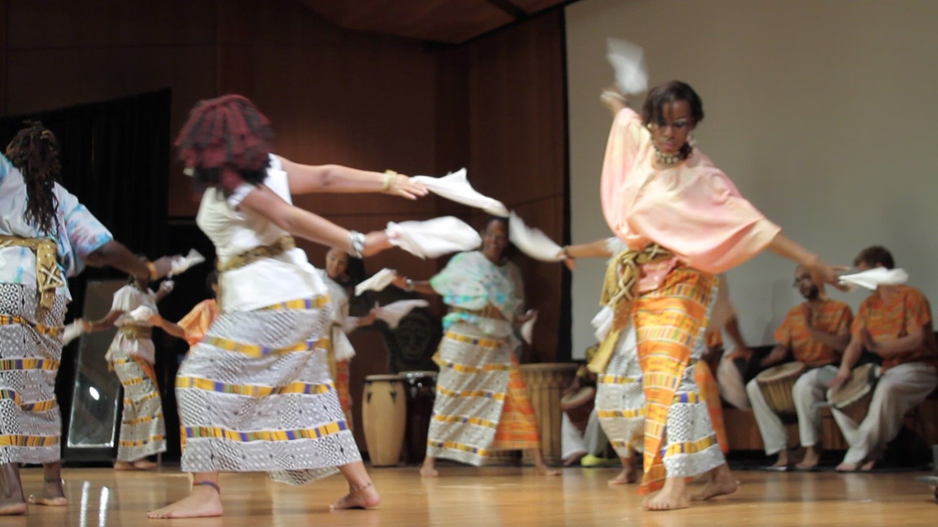 People dancing on a stage, dressed in African-style clothing, holding white cloths, with drummers in the background.