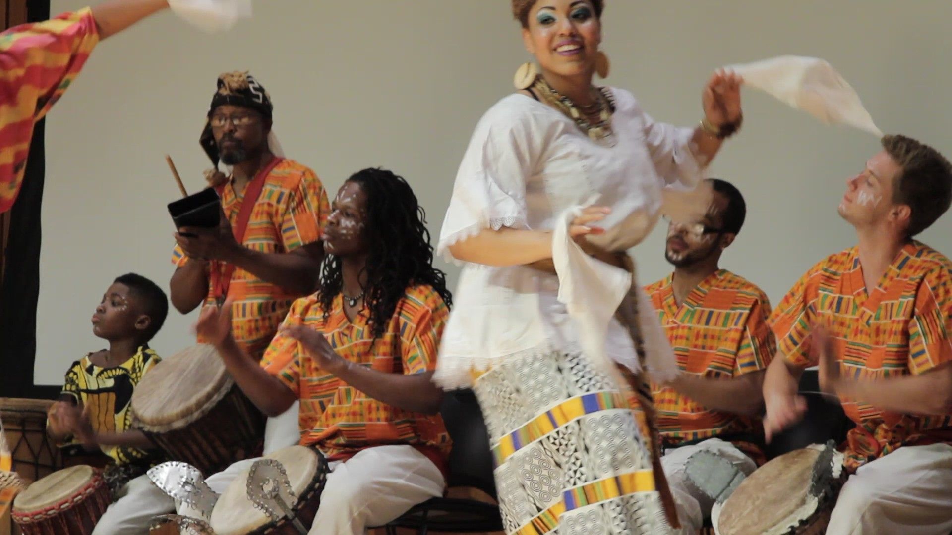 Group of musicians playing drums, woman dancing in white, colorful orange and gold clothing.