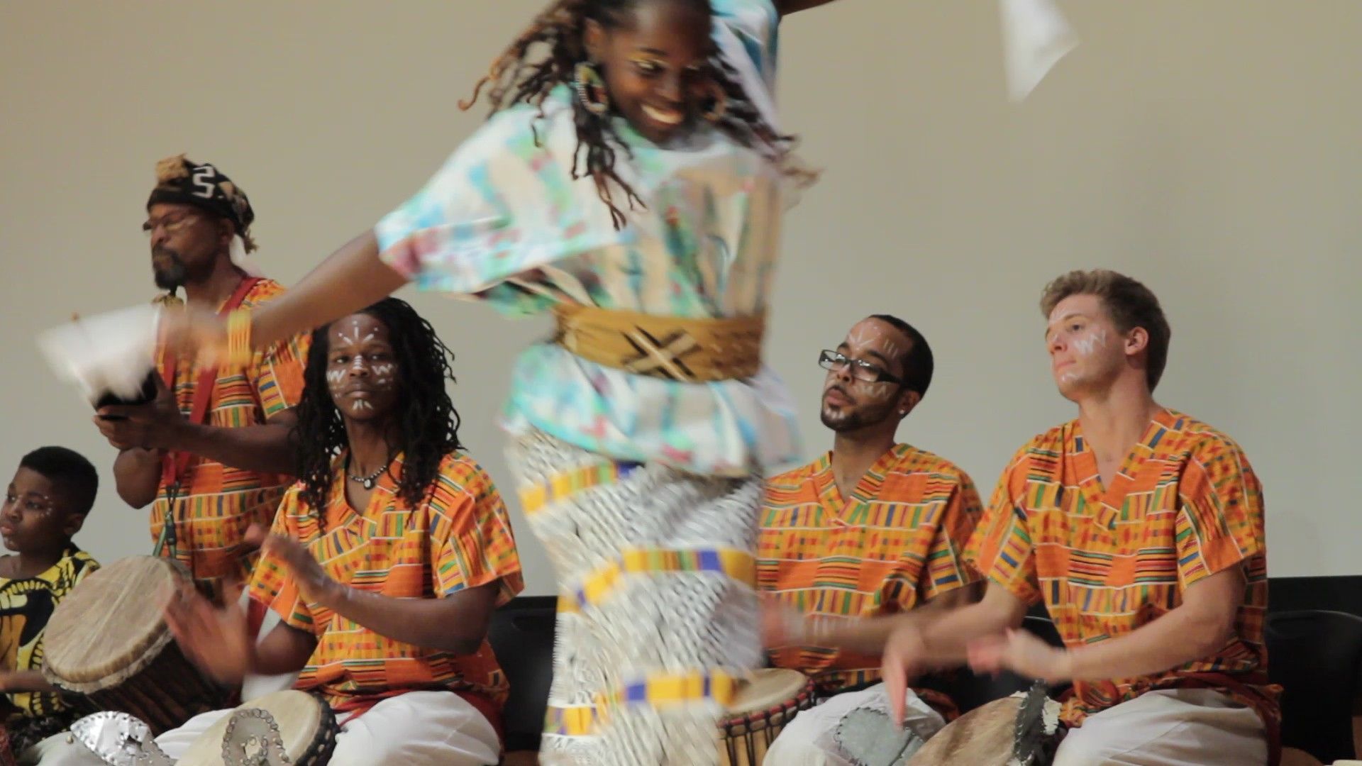A woman dances joyfully while a band plays drums; everyone wears colorful African-print clothing.