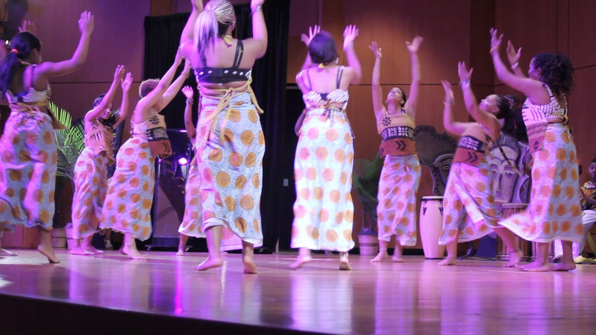 Group of women dancing on a stage, arms raised. They wear matching patterned dresses, lit by stage lights.