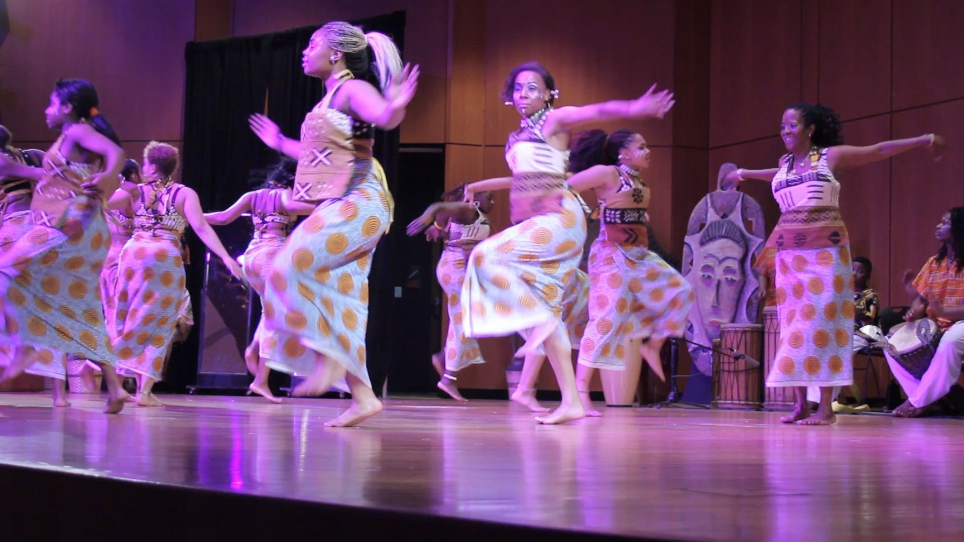 Dancers in colorful patterned dresses perform on a stage under purple lighting.