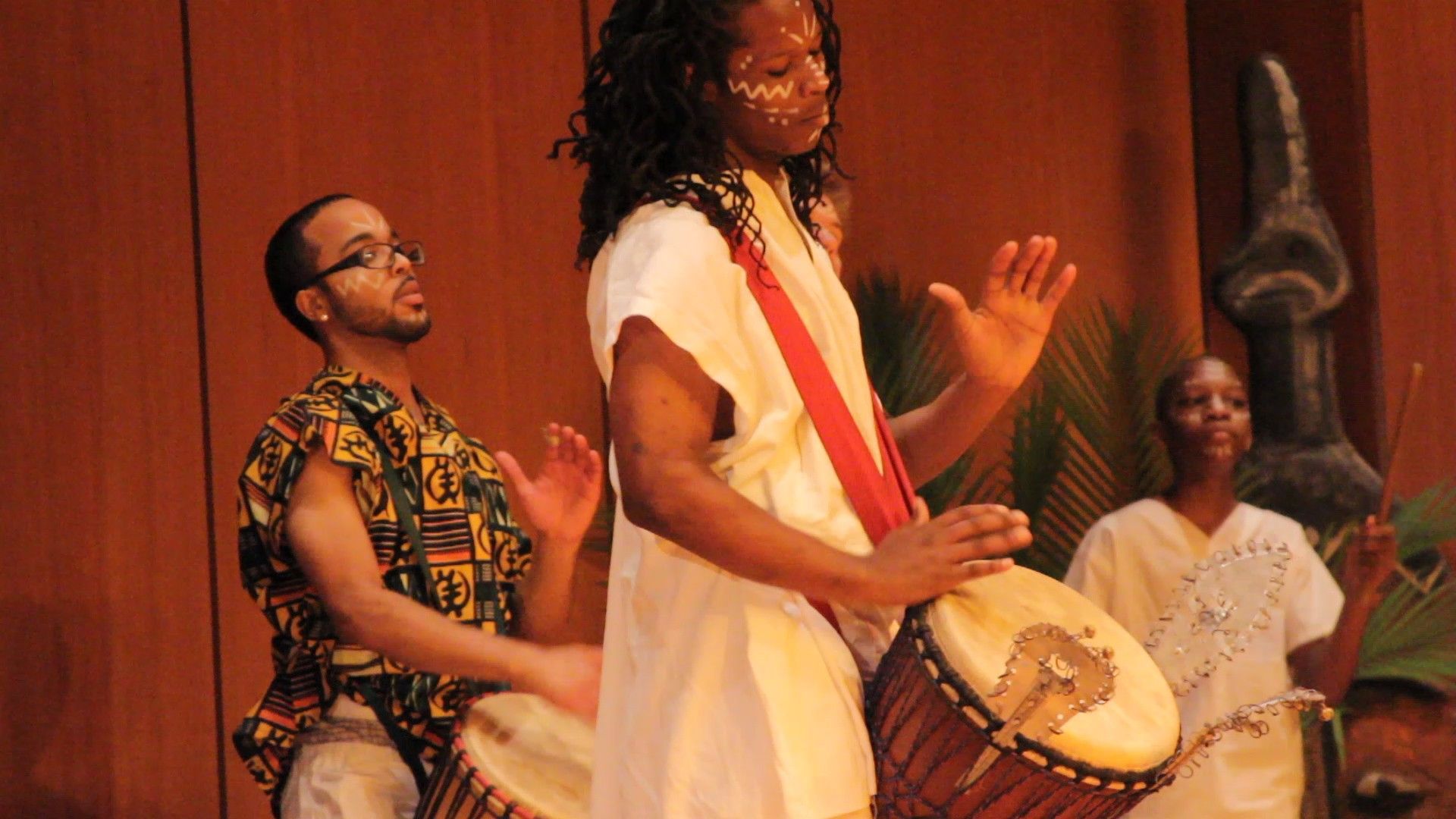 Three people playing drums on a stage, one with face paint, all in white and colorful clothing.