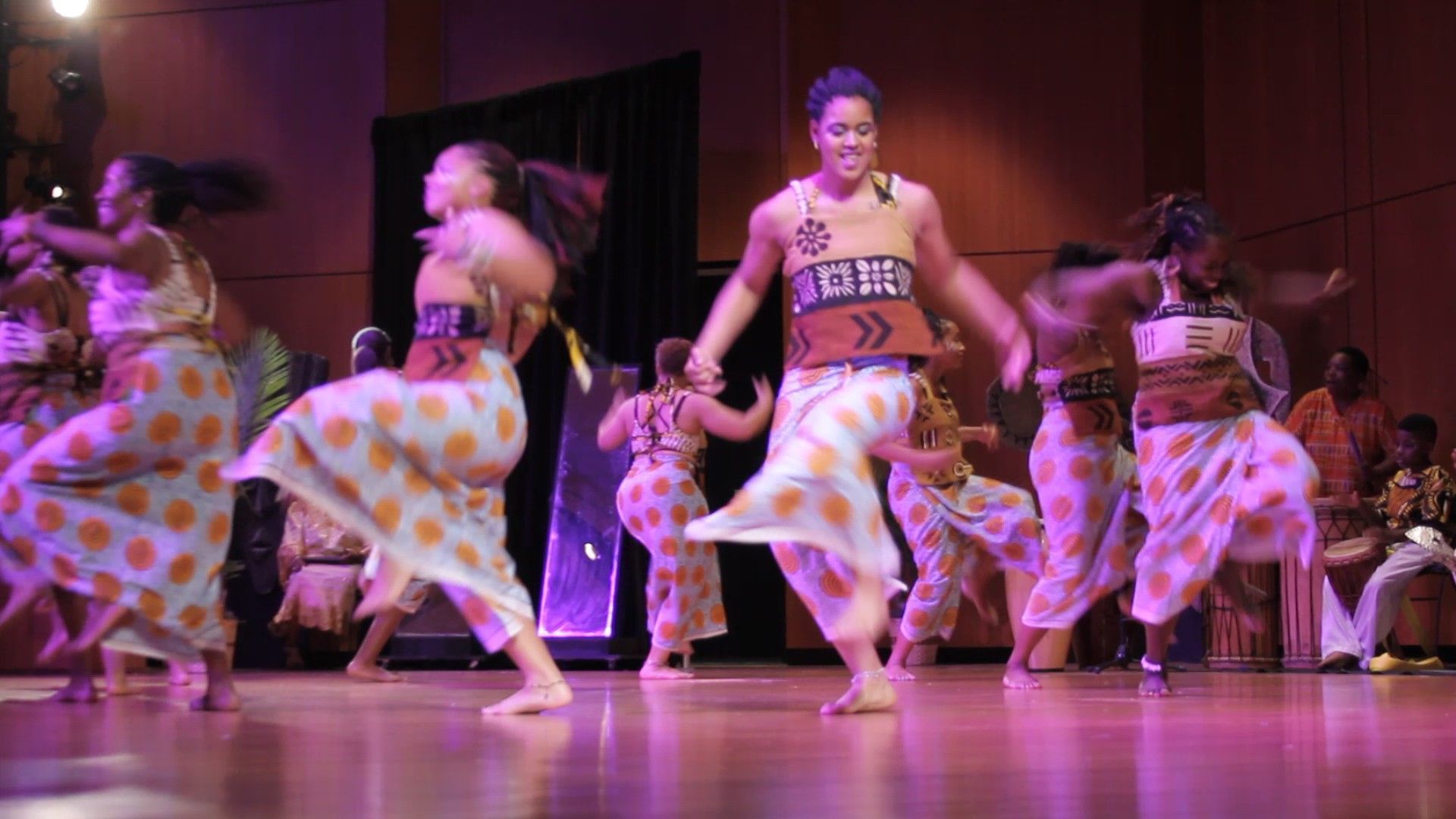 Dancers in patterned skirts and tops perform on stage with purple lighting.