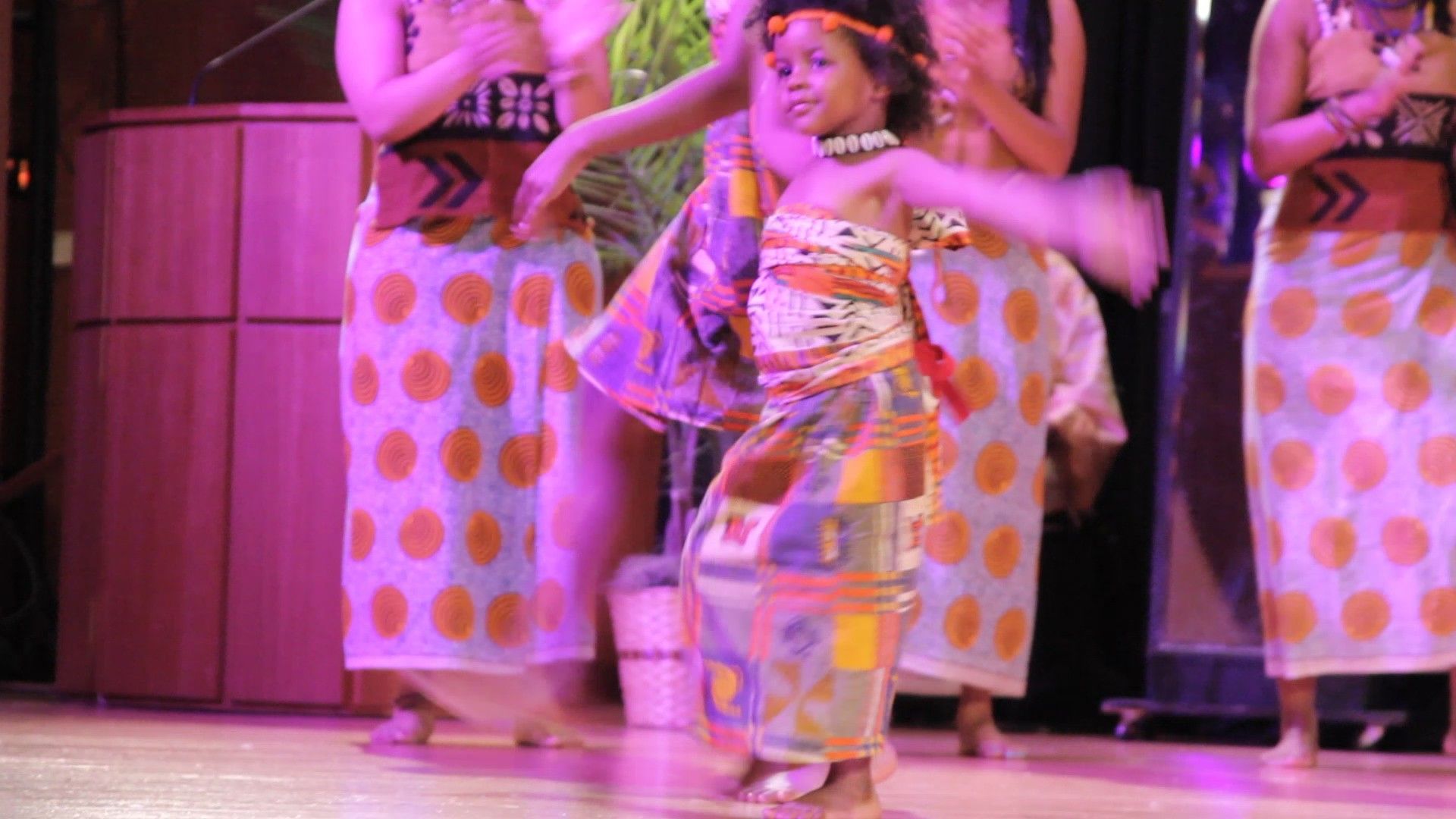 Girl in patterned outfit dances on stage with other dancers.