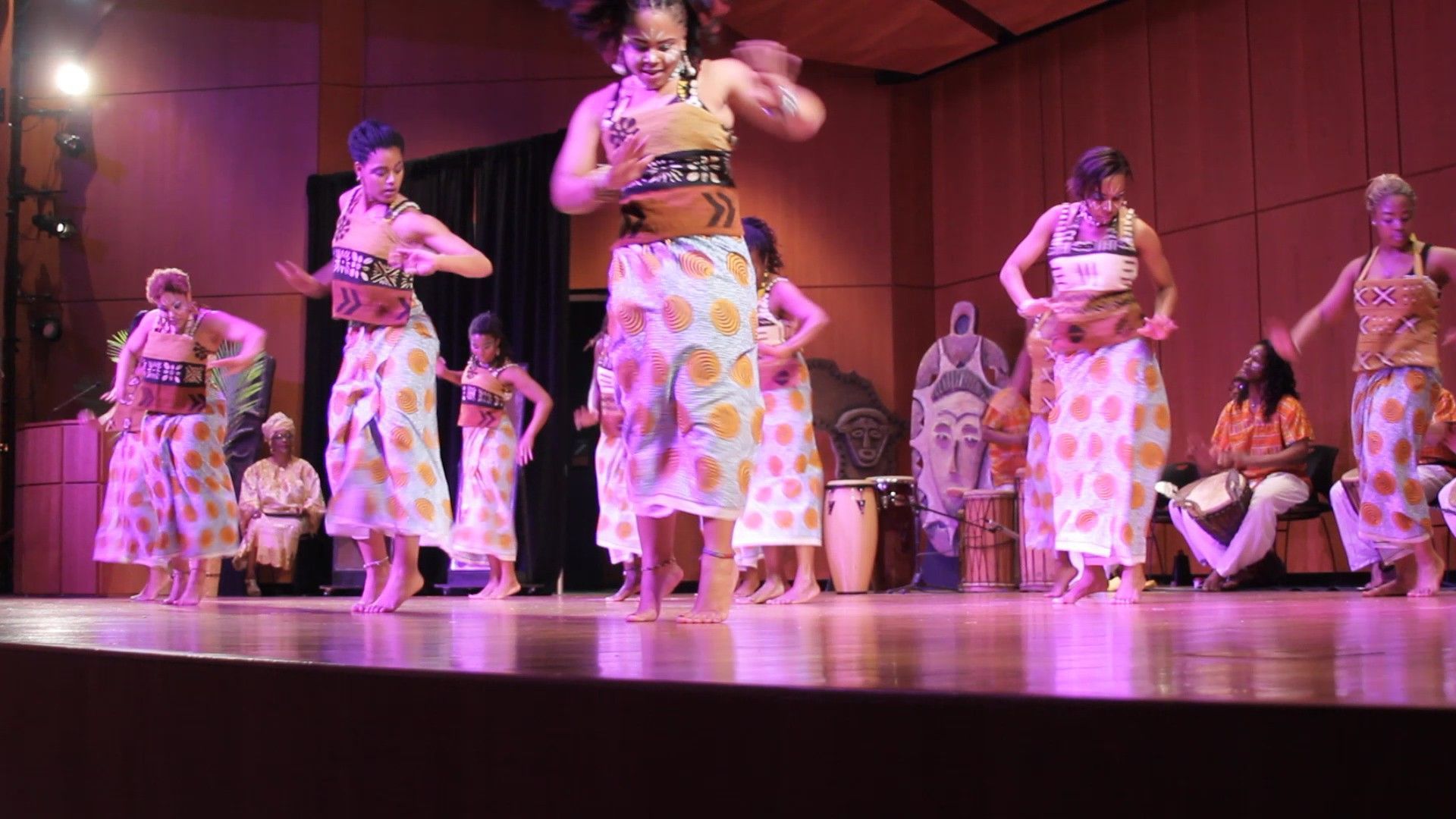 Dancers on stage in colorful outfits, performing. Musicians in background.