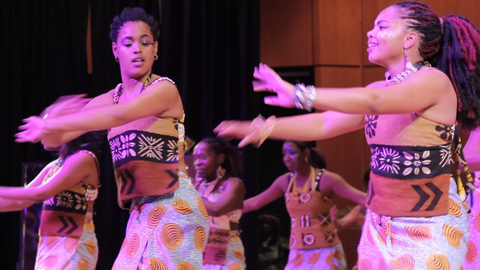 African dancers perform on stage. Women wear patterned skirts and tops, arms raised, under warm lighting.