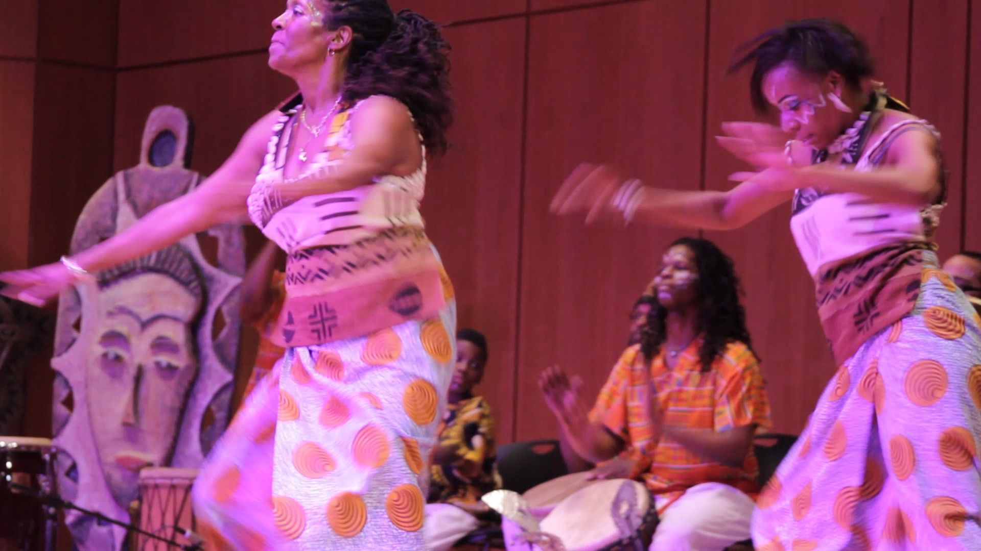 Two women in patterned skirts dance on stage, performers with drums behind them.