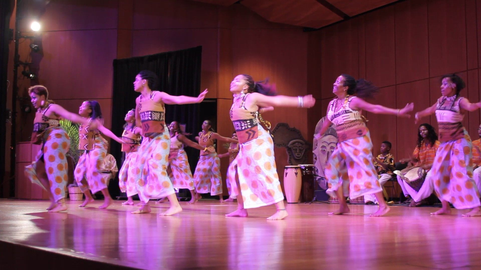 Dancers in colorful outfits perform on stage, arms outstretched, with a band playing drums in the background.