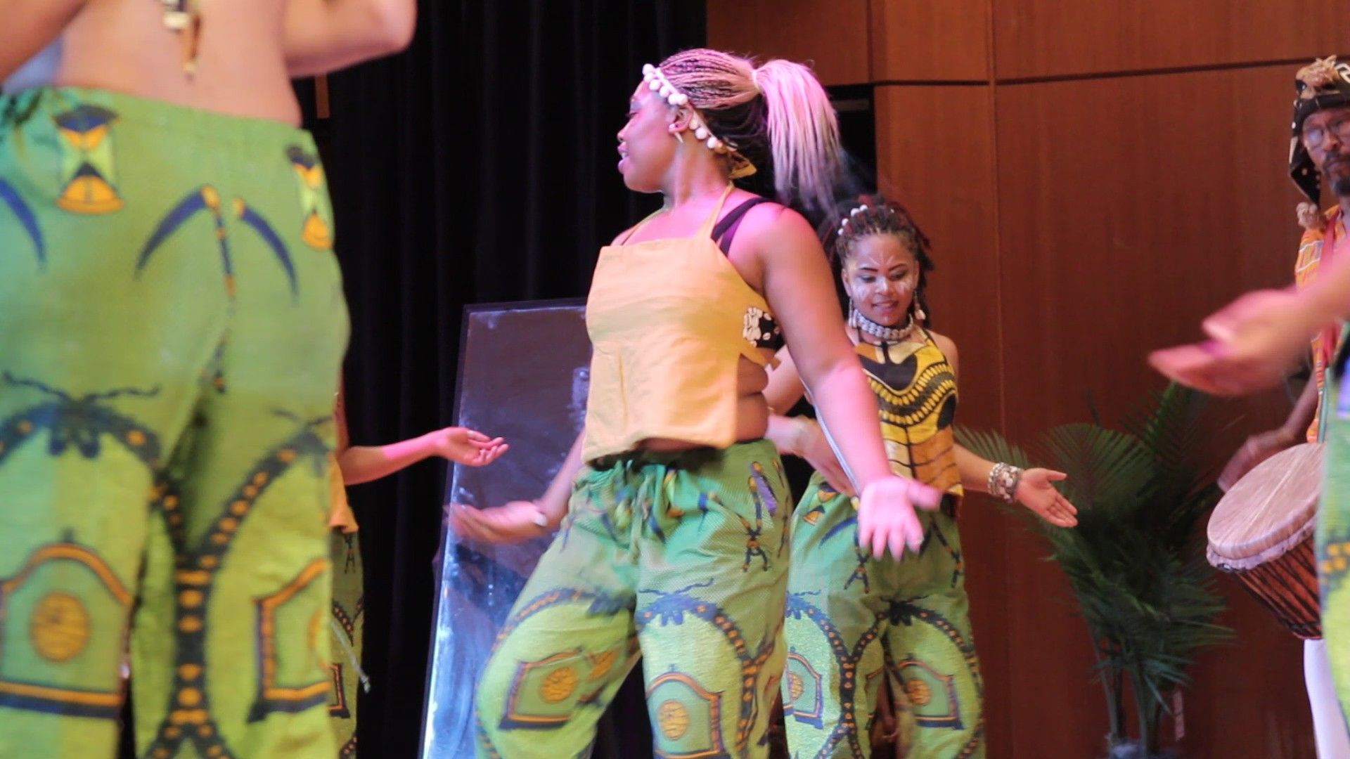Dancers in yellow tops and green patterned pants perform on stage.