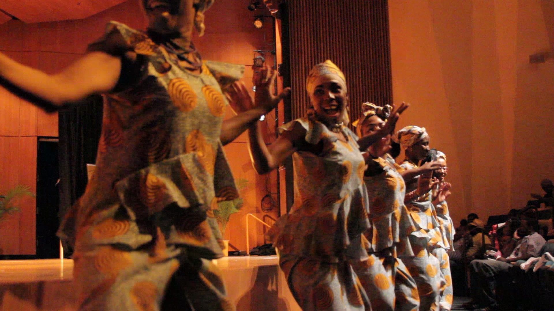Dancers in patterned dresses perform on stage; warm lighting.