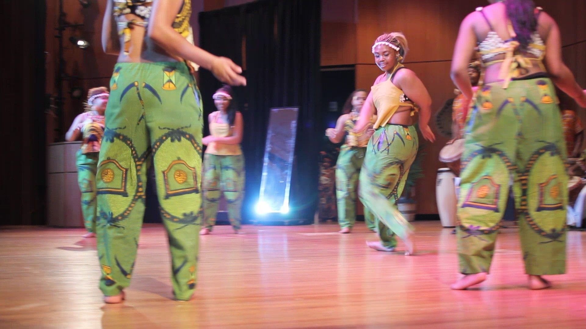 Dancers in green patterned pants and yellow tops perform on a wooden stage.