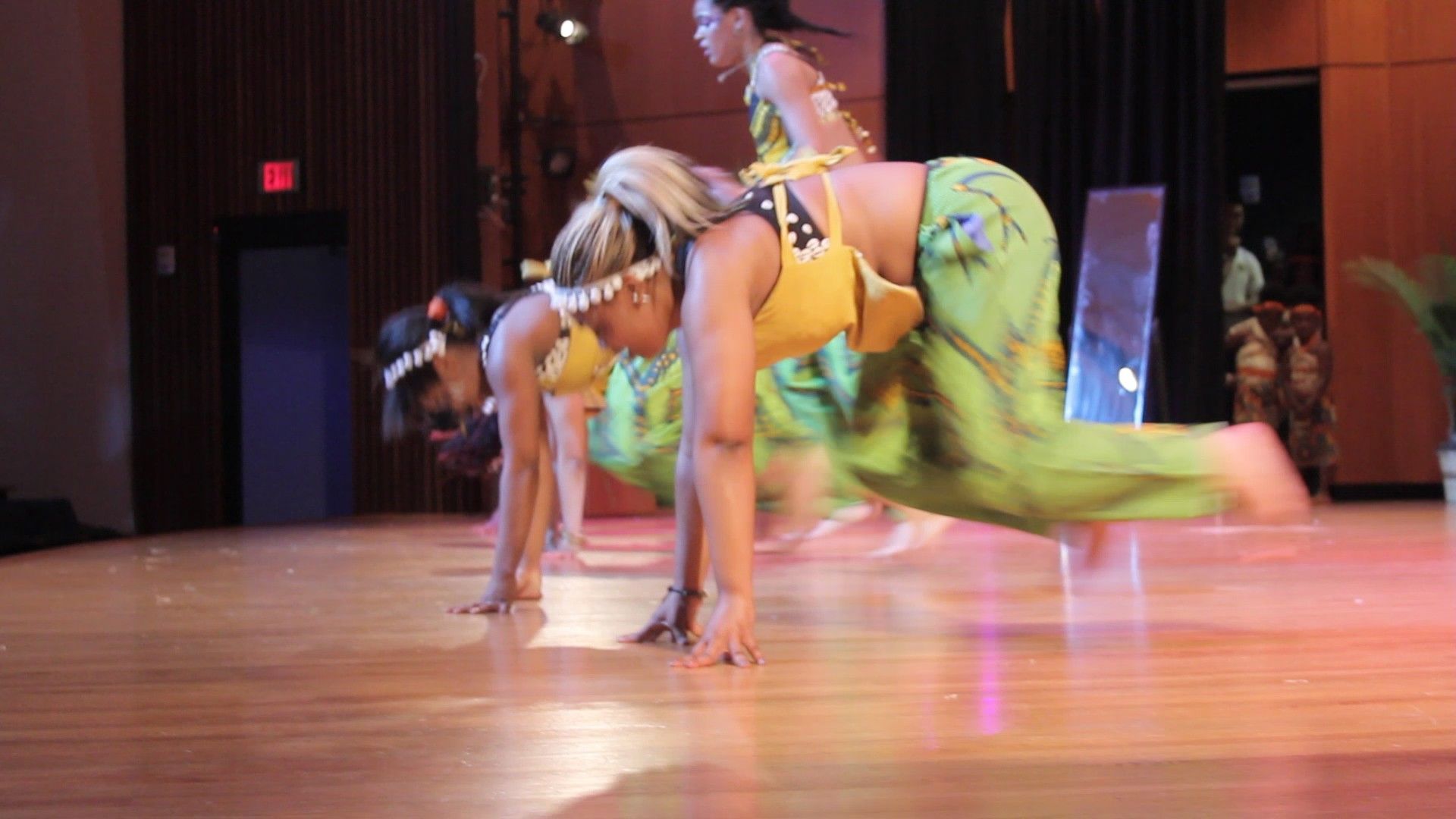 Dancers in vibrant outfits perform a low crawl on stage, illuminated by colorful lights.