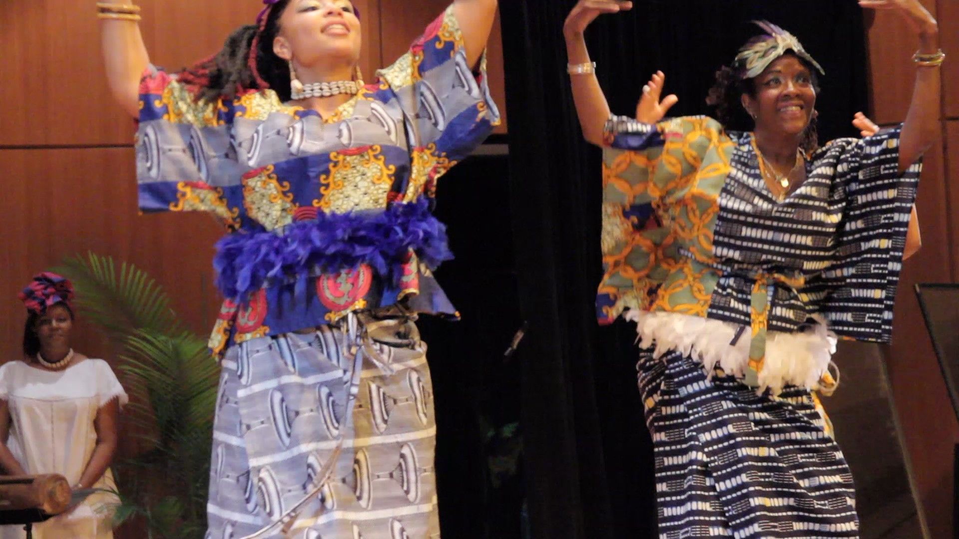 Two women in African attire dance onstage with arms raised, smiling.