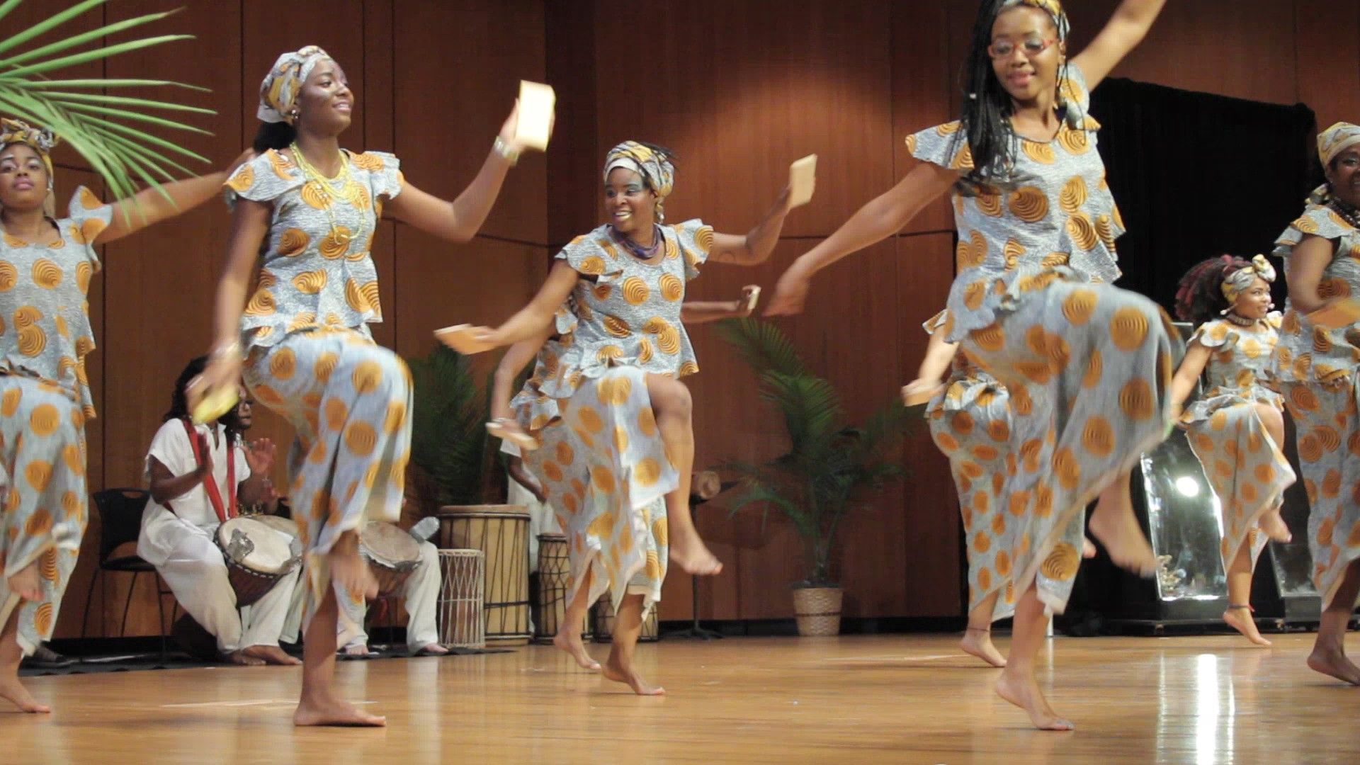 Women in matching blue and yellow outfits dance on a stage with a band playing drums and instruments.