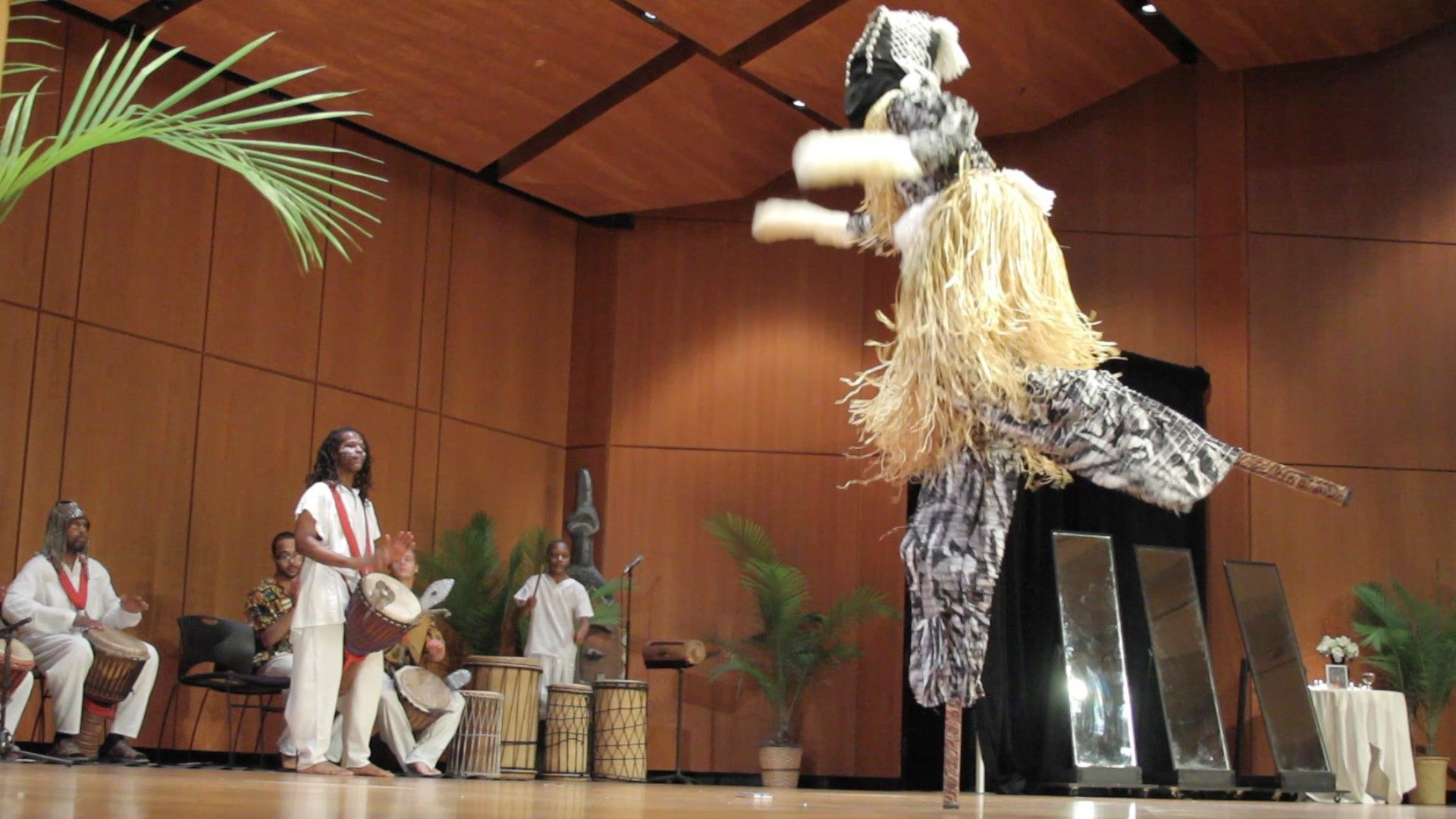 Performance with a large, tall mask and musicians on stage in a wood-paneled hall.