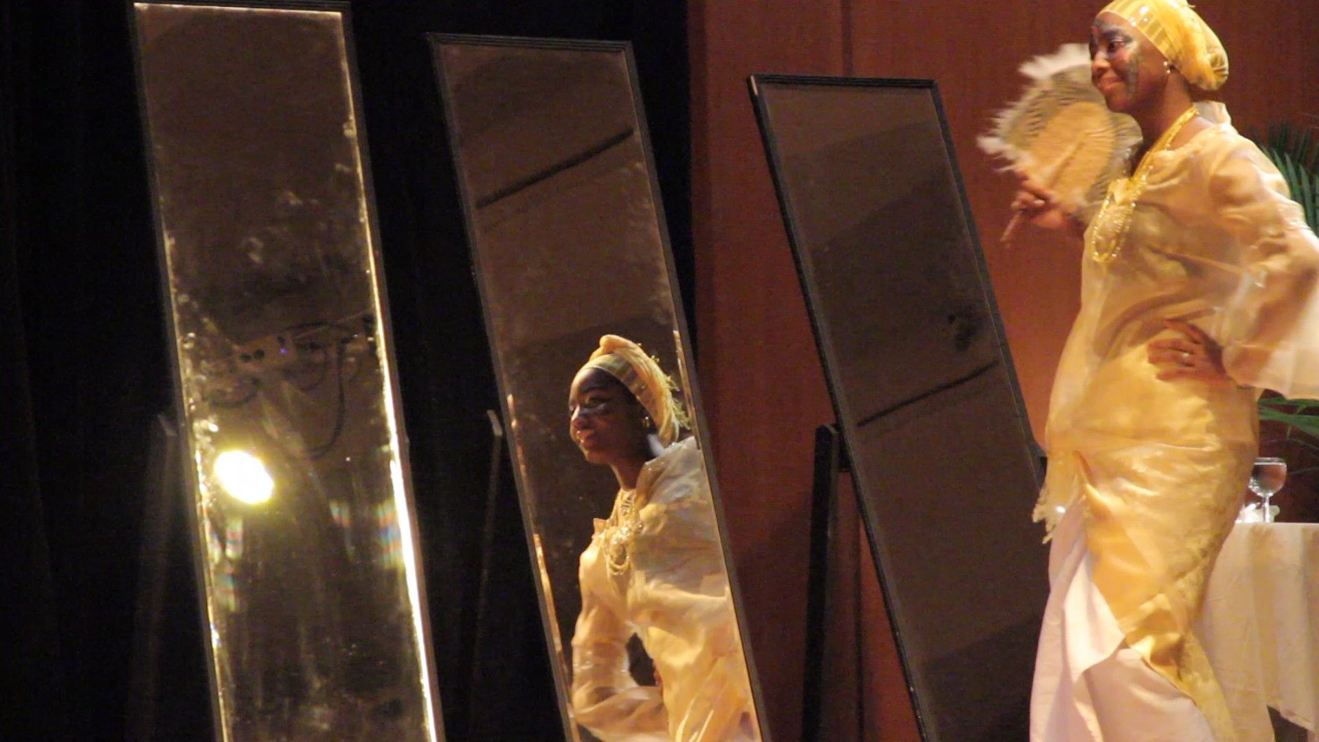 Woman in yellow outfit poses in front of three mirrors on a stage, holding a fan.