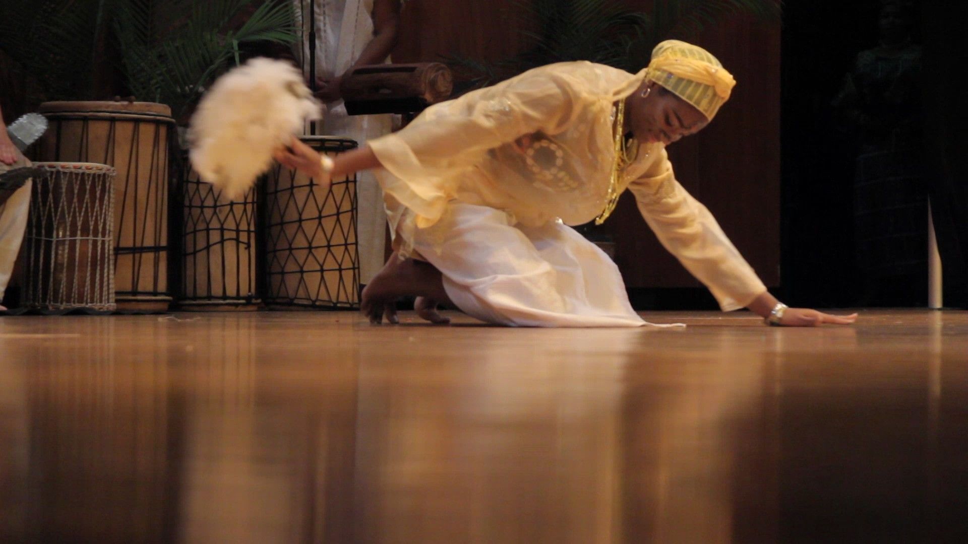 Person in yellow kneeling, holding a fan, on a stage with drums in the background.