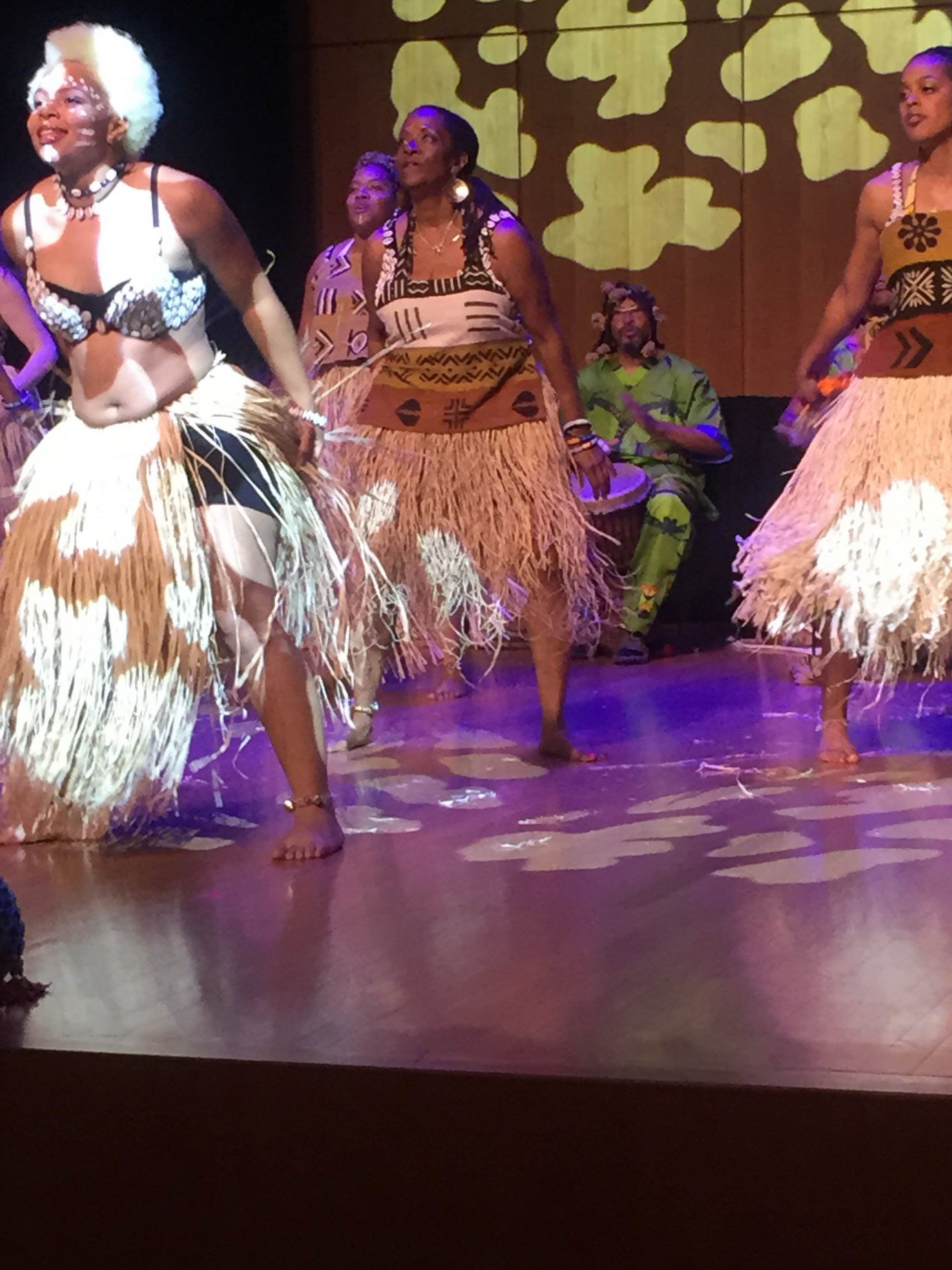 Dancers in tribal costumes performing on stage. Musicians in background. Stage lit with spotlight.