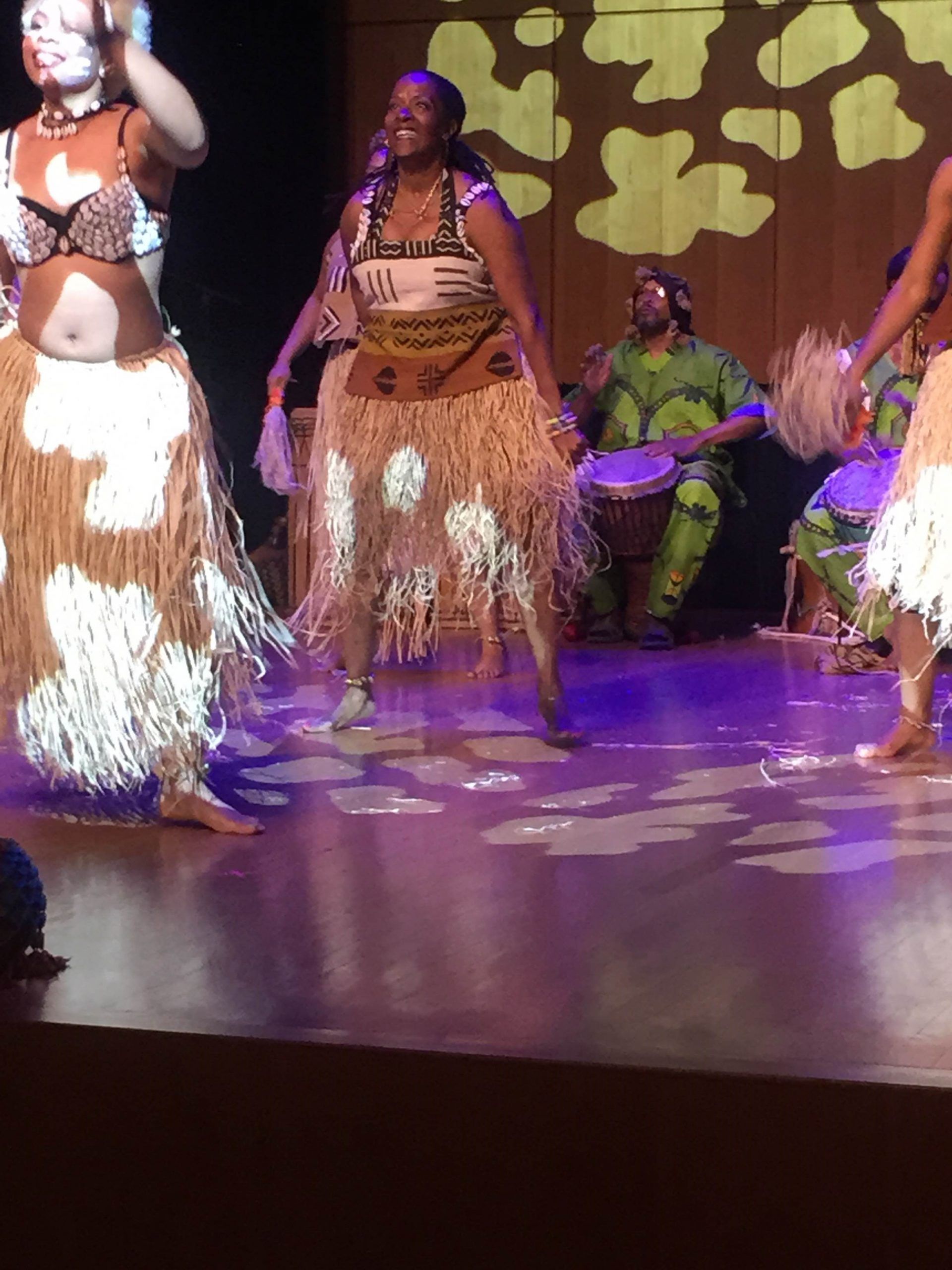 African dancers perform on stage, wearing tribal-inspired outfits. A drummer in green sits behind them.