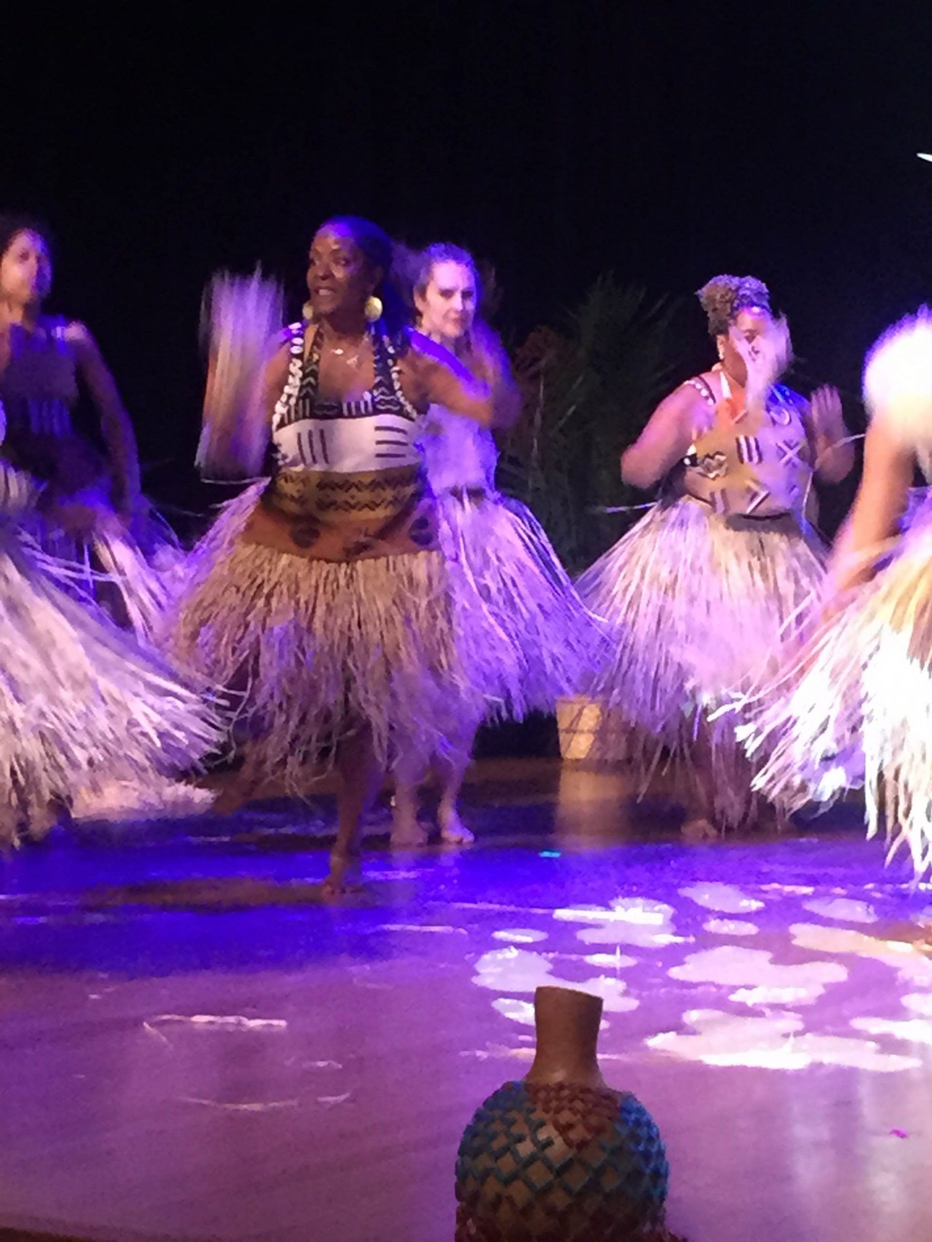 Women performing a traditional dance on a stage; wearing grass skirts and tops with tribal patterns, lit by blue light.