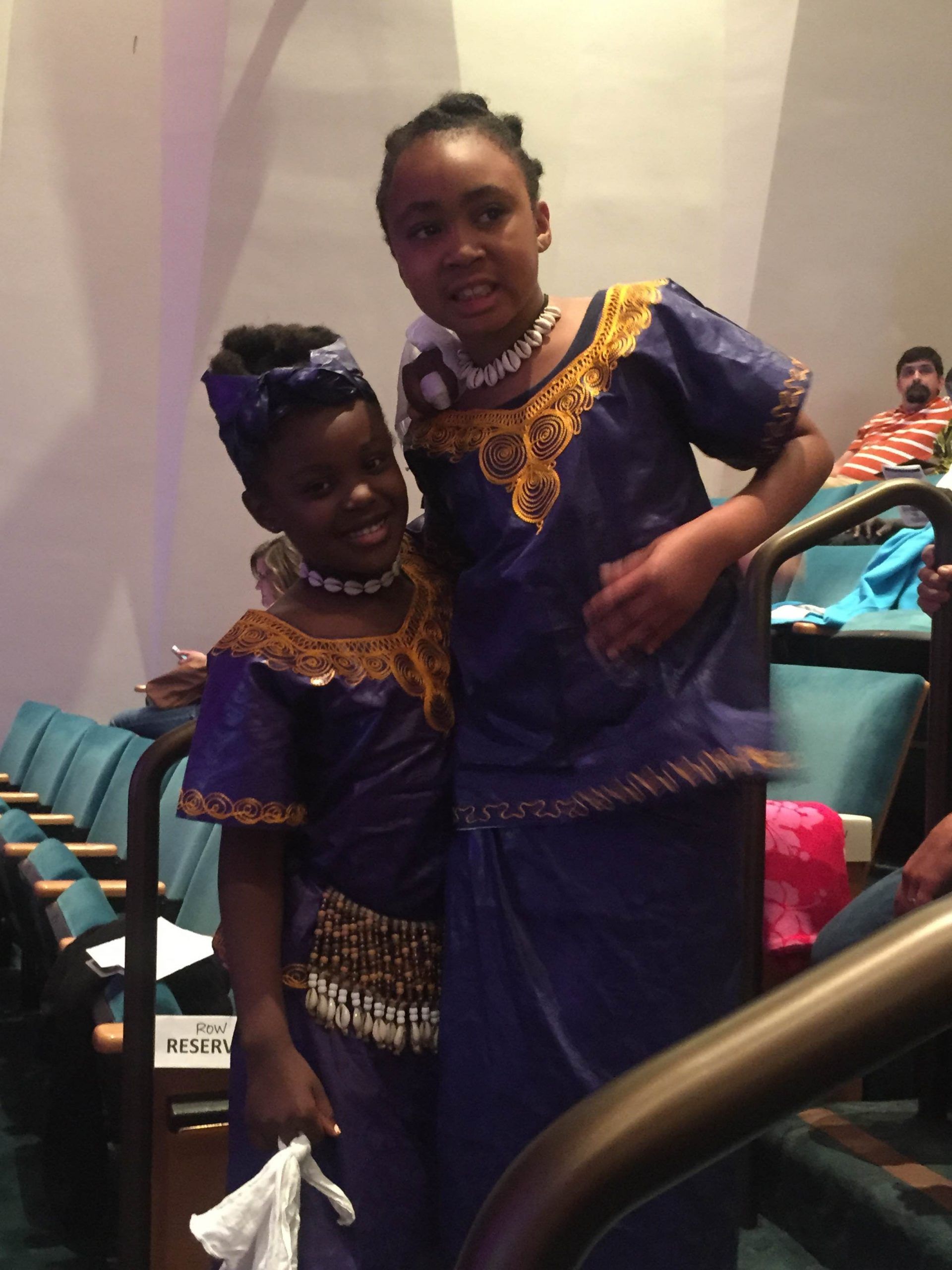 Two young girls in blue and gold traditional outfits, posing inside a theater.