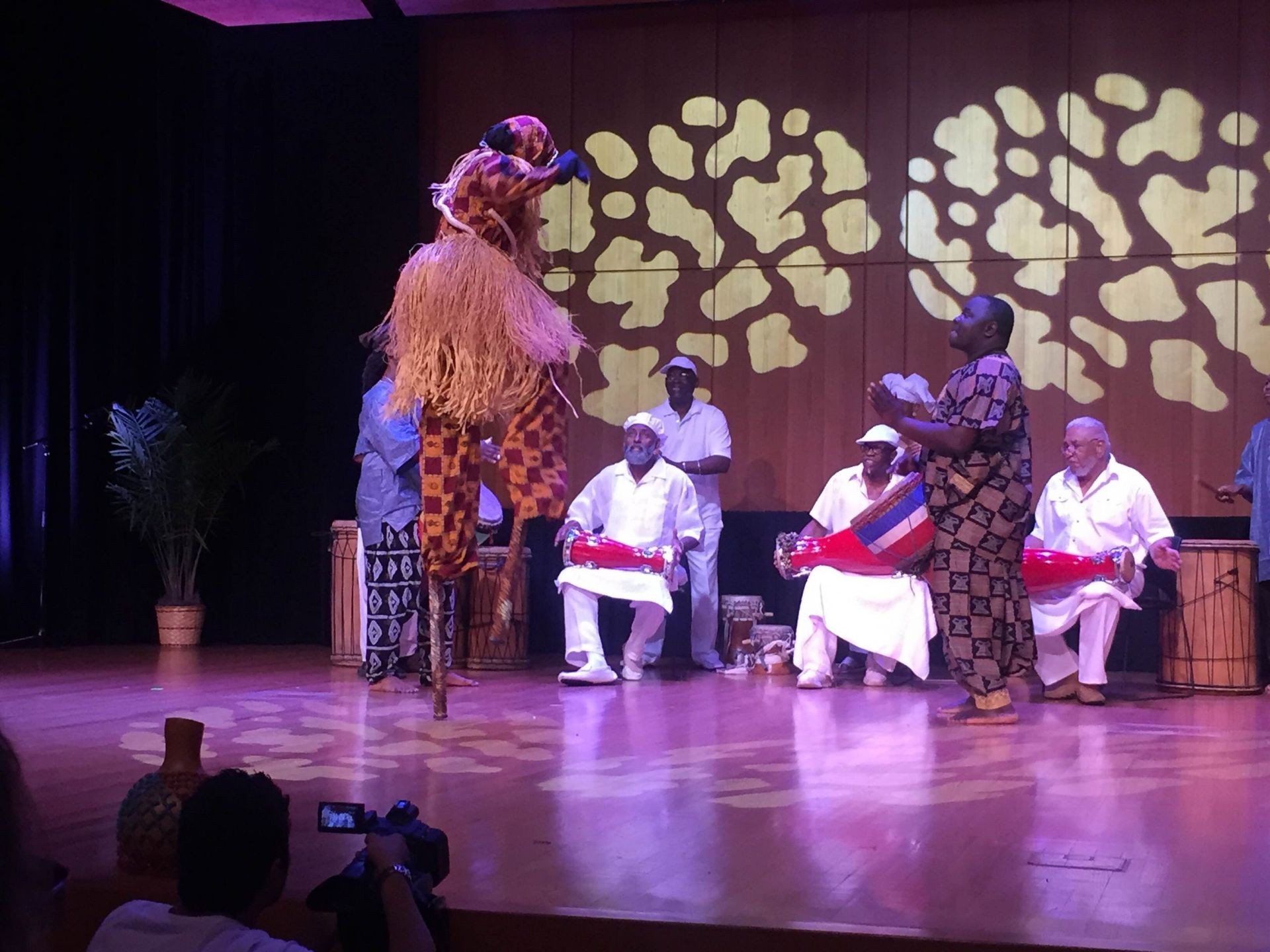 African stilt walker performs on stage with musicians; warm lighting, patterned backdrop.