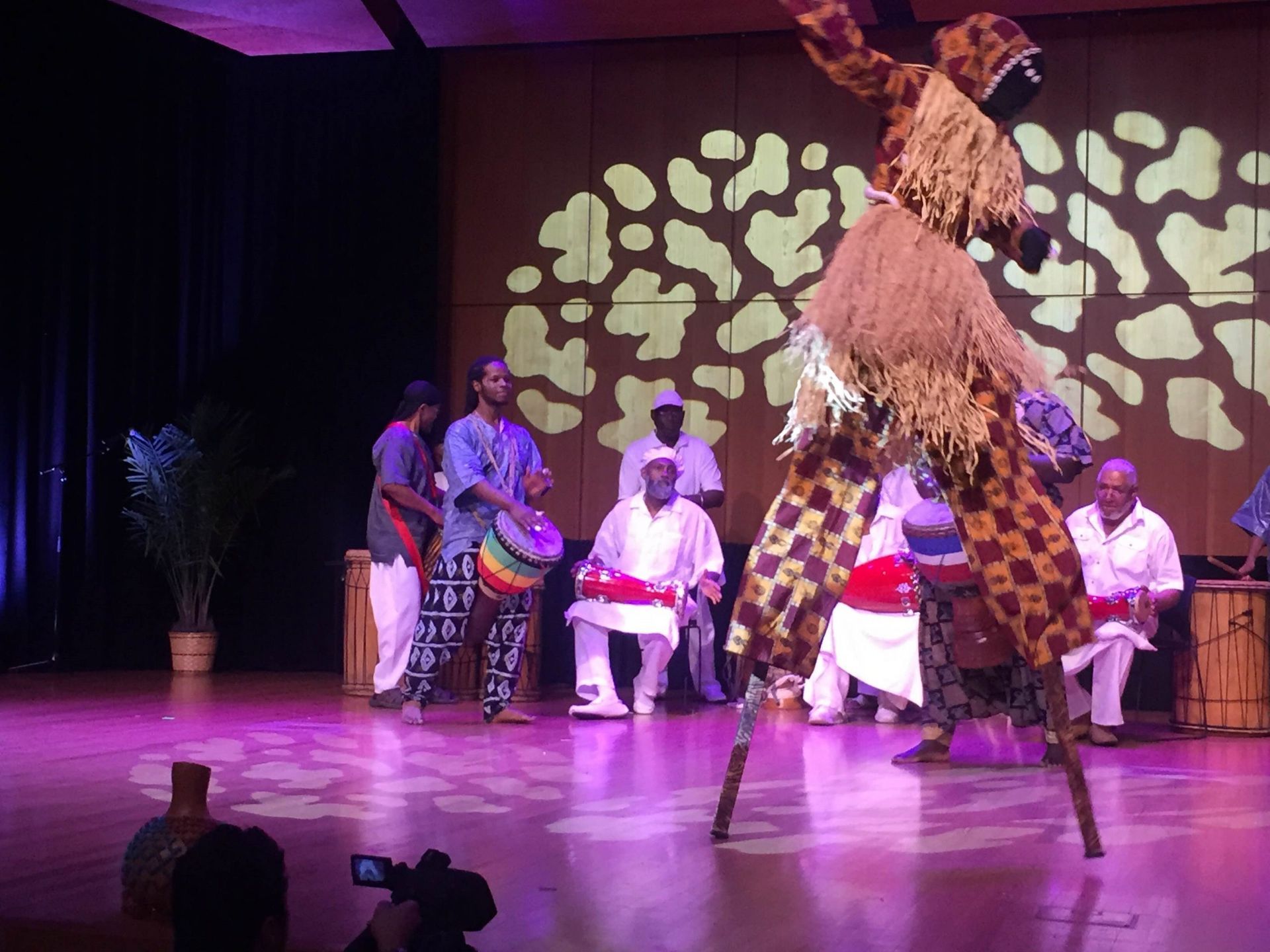 African dancers and musicians on a stage. A stilt walker performs as a drum ensemble plays.