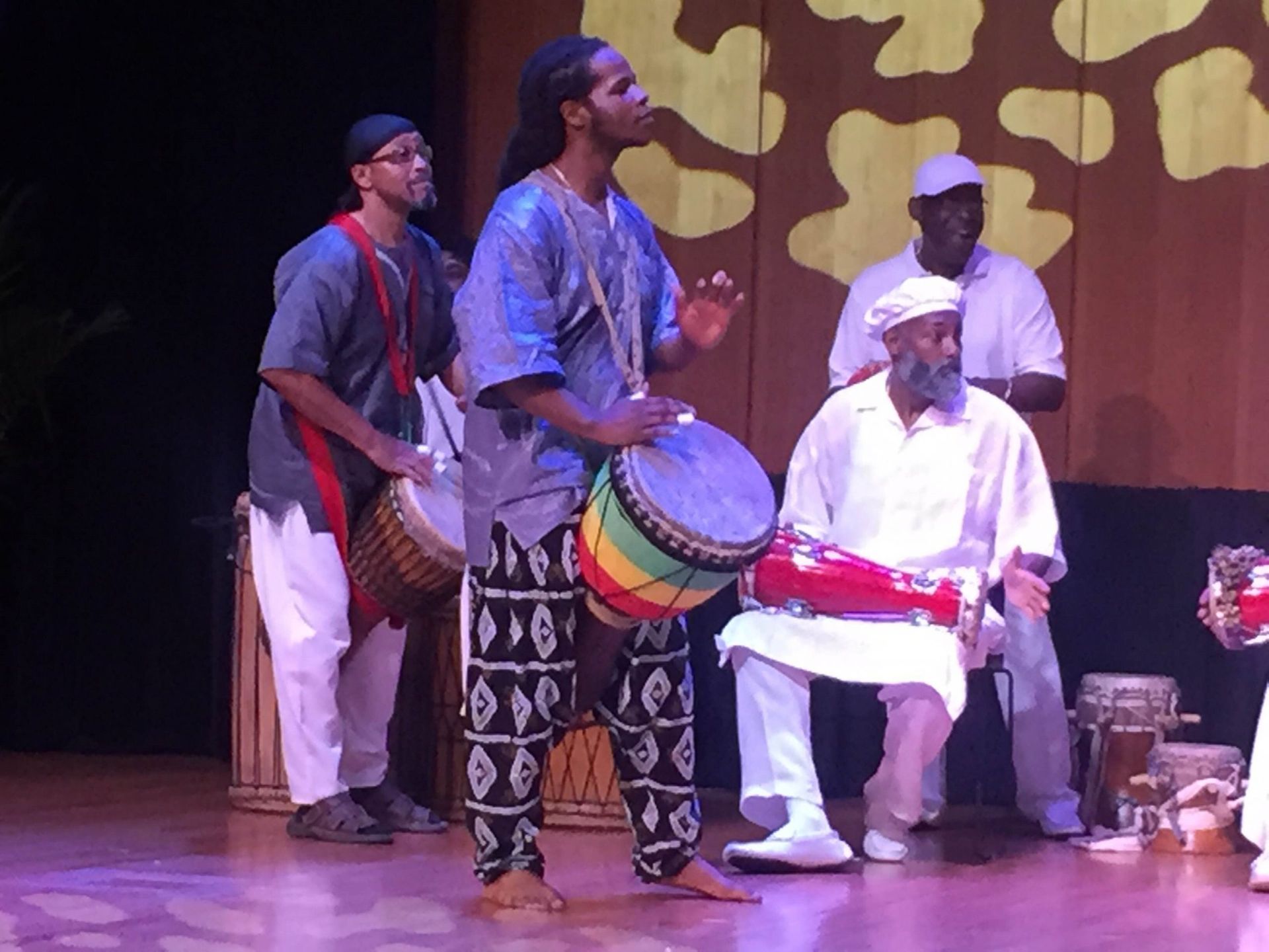 A band performs on stage, playing drums. Musicians wear colorful attire.