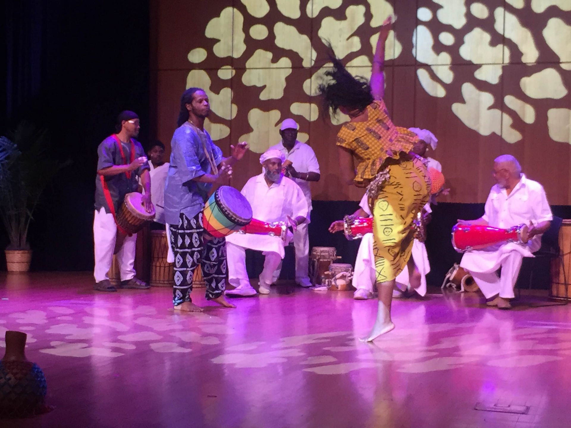 Dancers and drummers on stage with patterned lighting. The dancer is mid-air in a colorful dress.