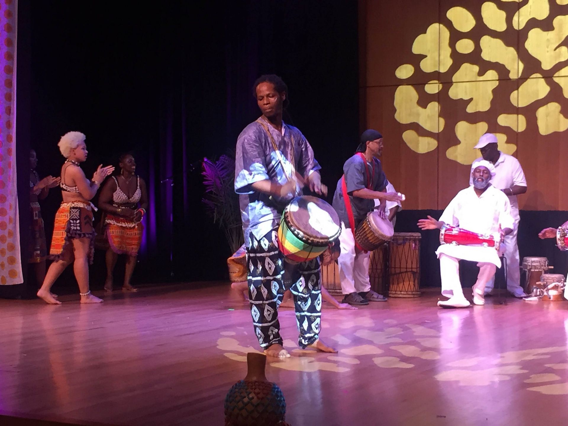 A group of people on stage performing African music; drummers and dancers on a wooden floor, lit by stage lights.