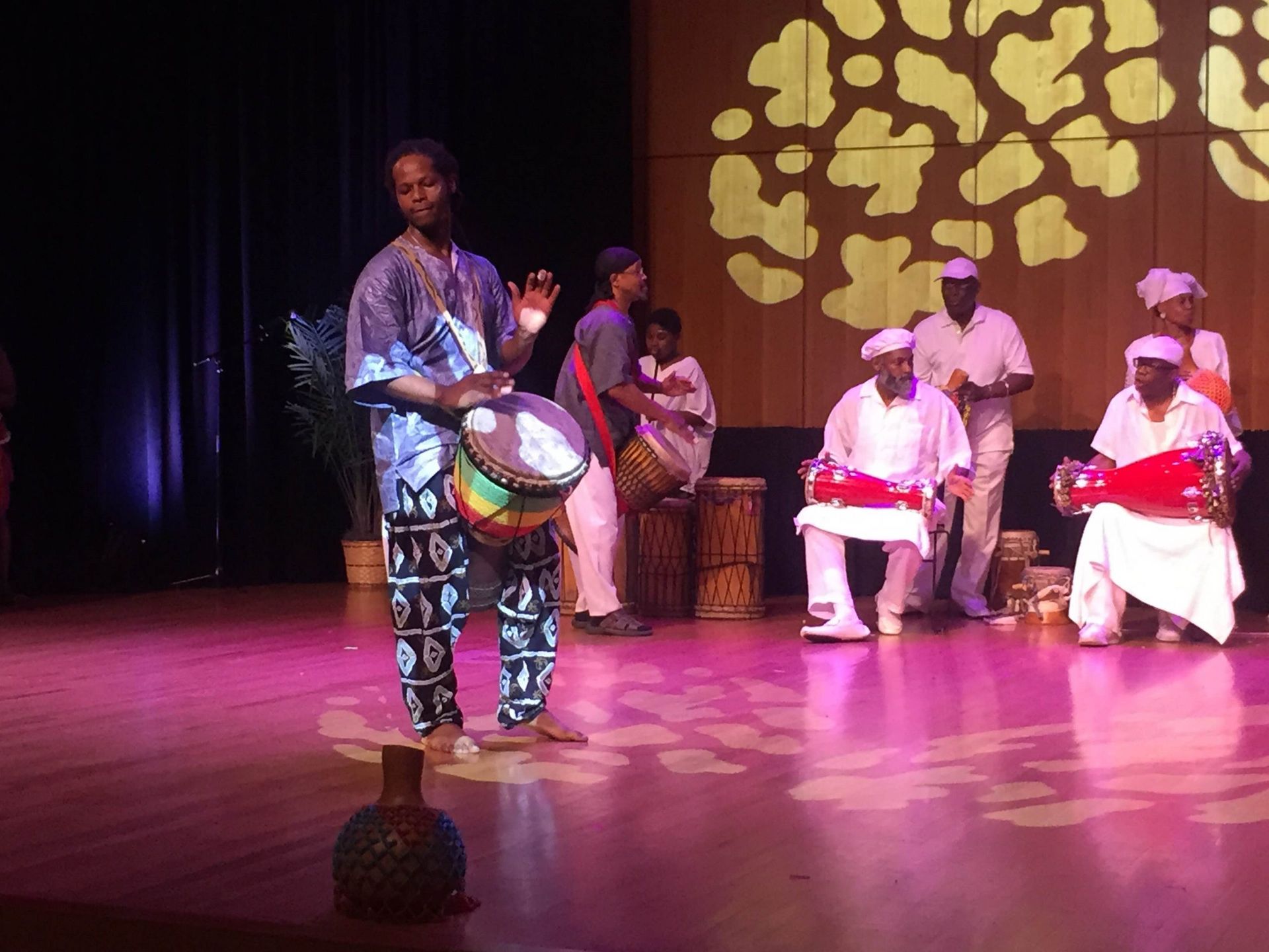 A man drumming on stage, flanked by musicians in white and others playing drums, under stage lights.