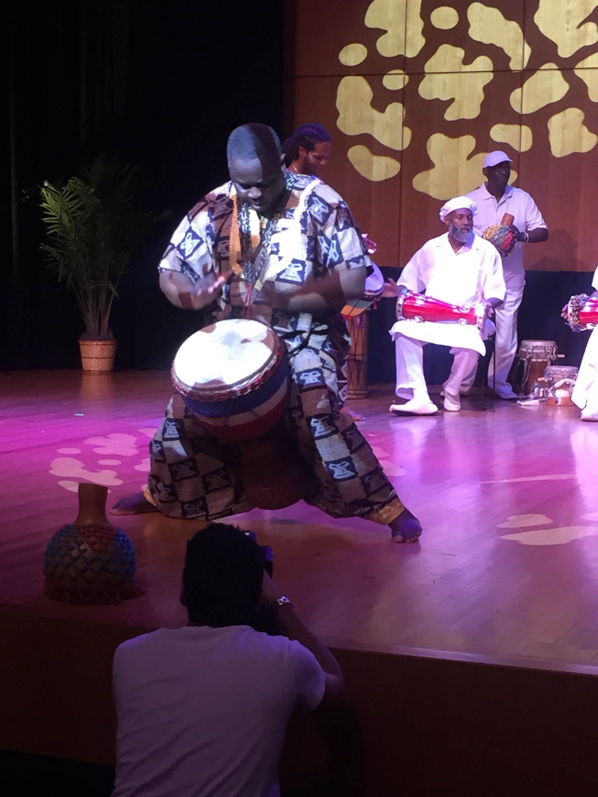 African drummer performing on stage, wearing patterned clothing. Audience member in foreground.