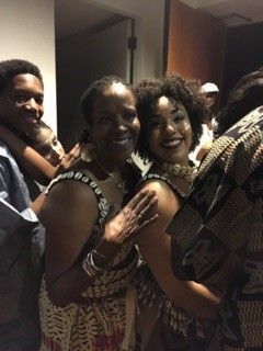 Two smiling women in patterned dresses embrace; people in background.