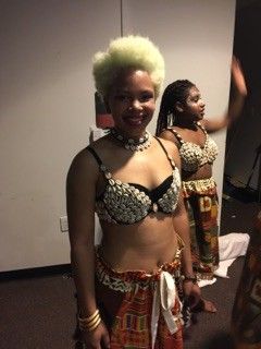 Woman with blonde afro in a decorative bra and skirt, smiling. Another woman in similar attire dances in the background.