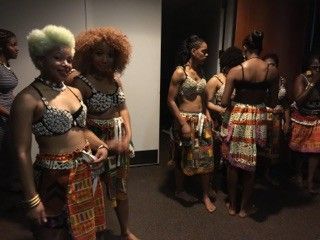Women in tribal outfits, backstage, preparing for a performance. Some wearing bedazzled bras and patterned skirts.
