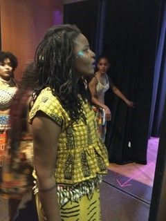 Woman in yellow African print outfit, backstage, talking. Other dancers in background.