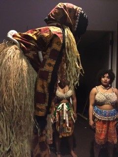A costumed figure with a long, grass-like skirt stands next to two women wearing tribal-style tops and skirts.