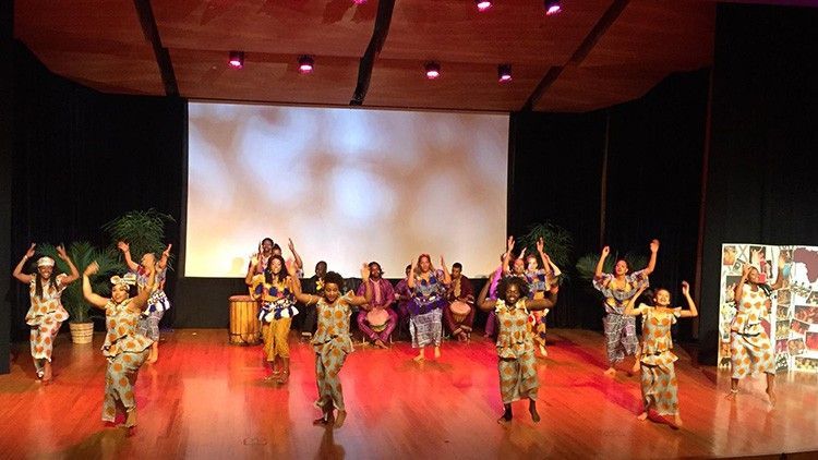 African dancers perform on a stage with drummers, colorful costumes, raising arms.
